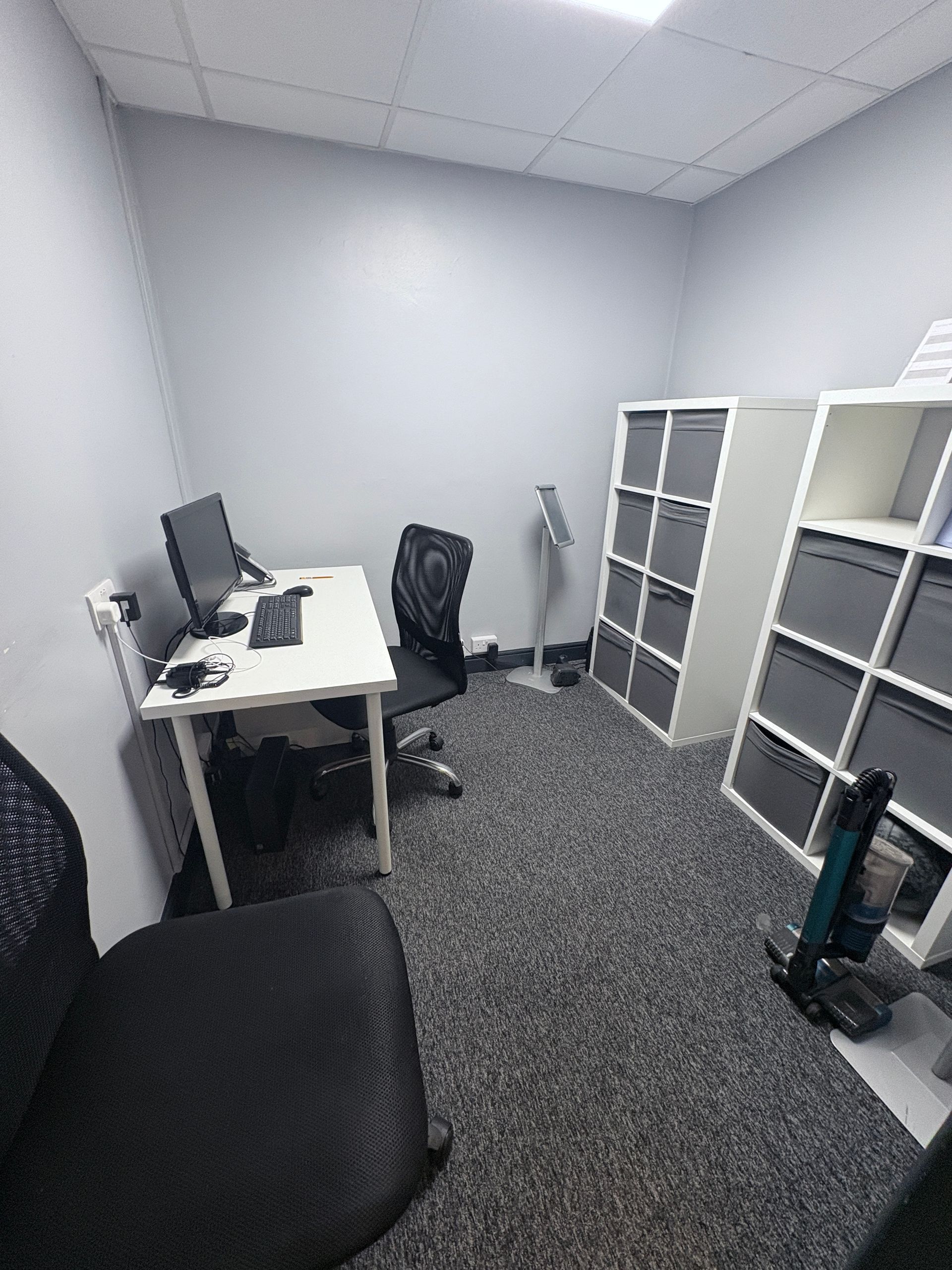Small office with a white desk, computer, black chair, and storage shelves with grey bins.