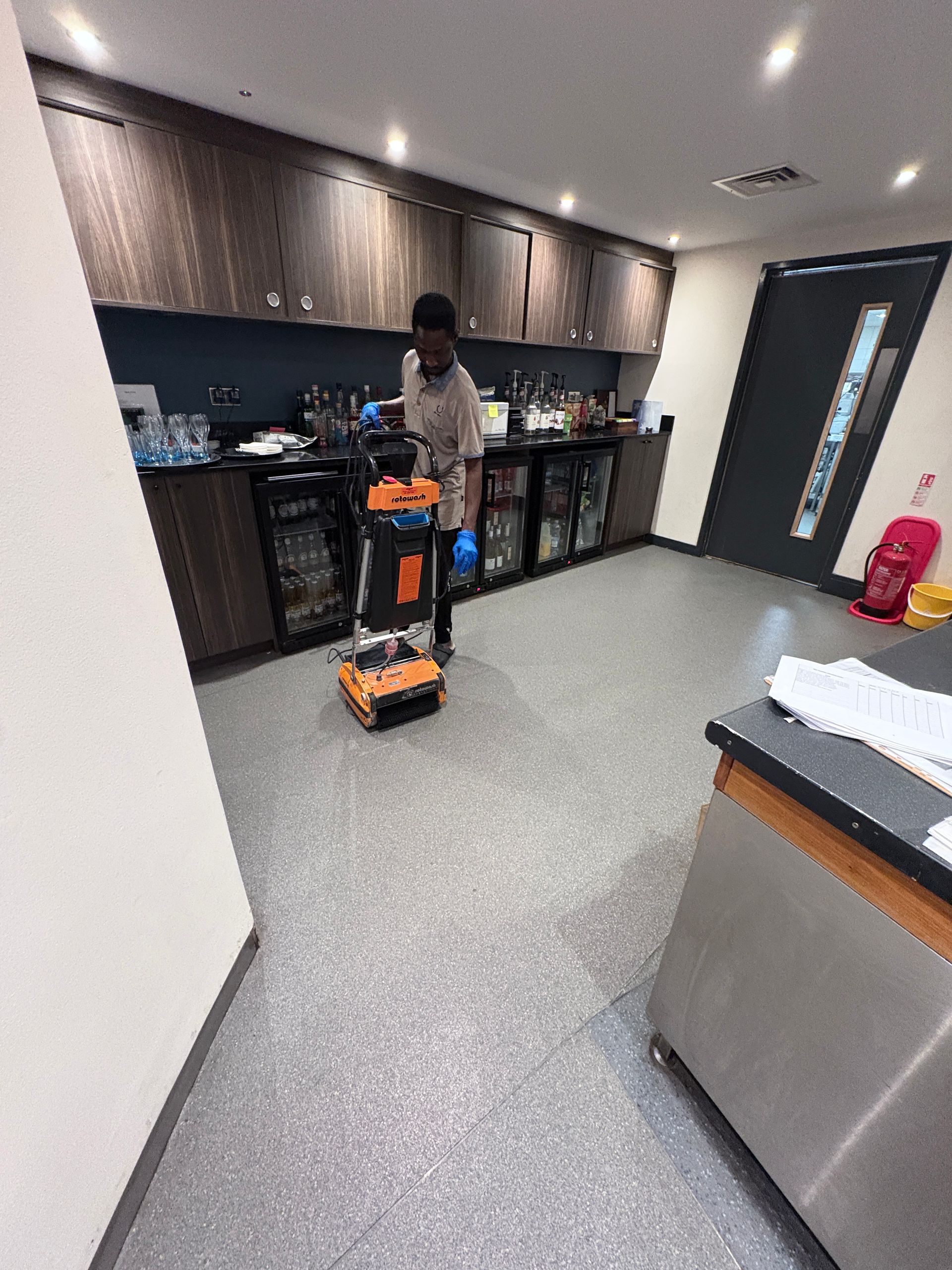 Person using a cleaning machine in a kitchen with gray floor, wood cabinets, and black appliances.