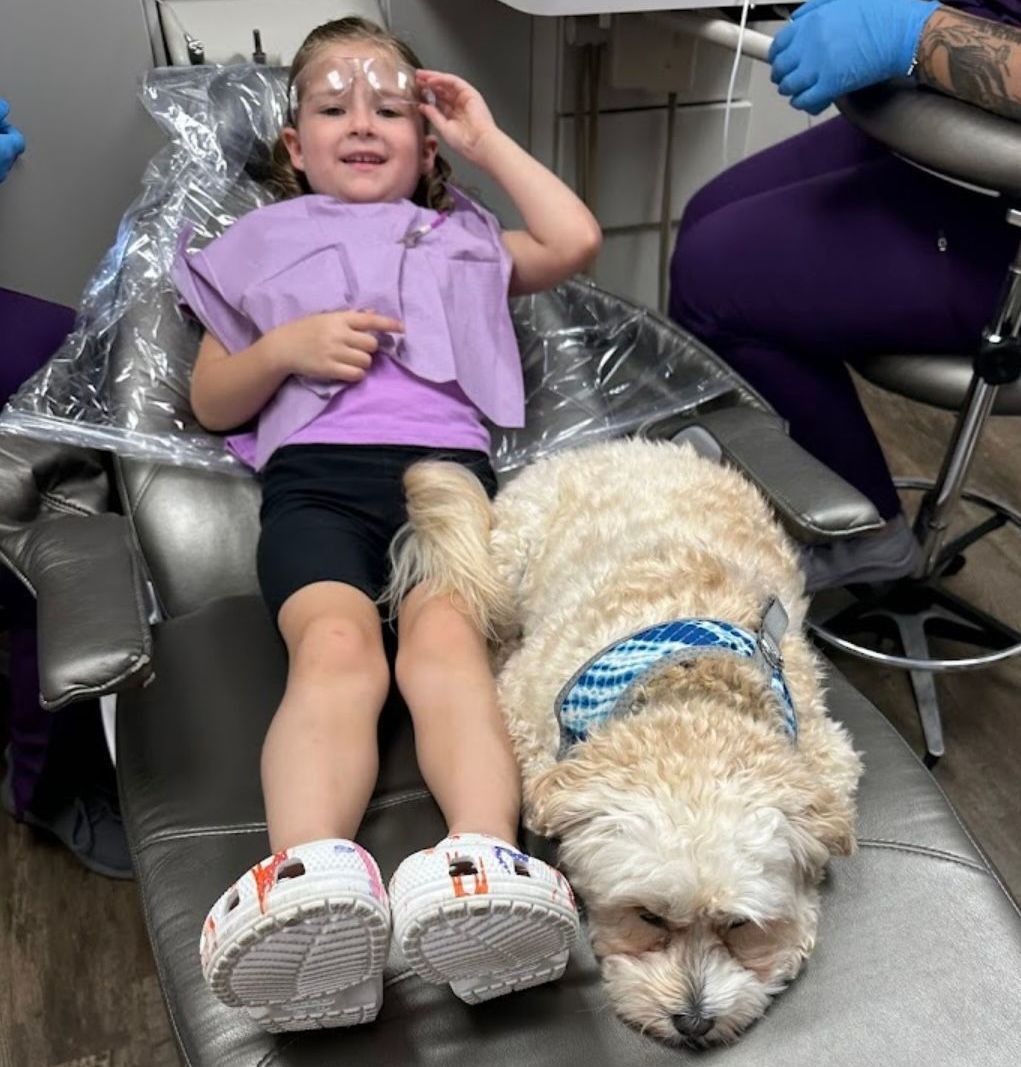 A little girl is sitting in a dental chair with a dental anxiety dog. 