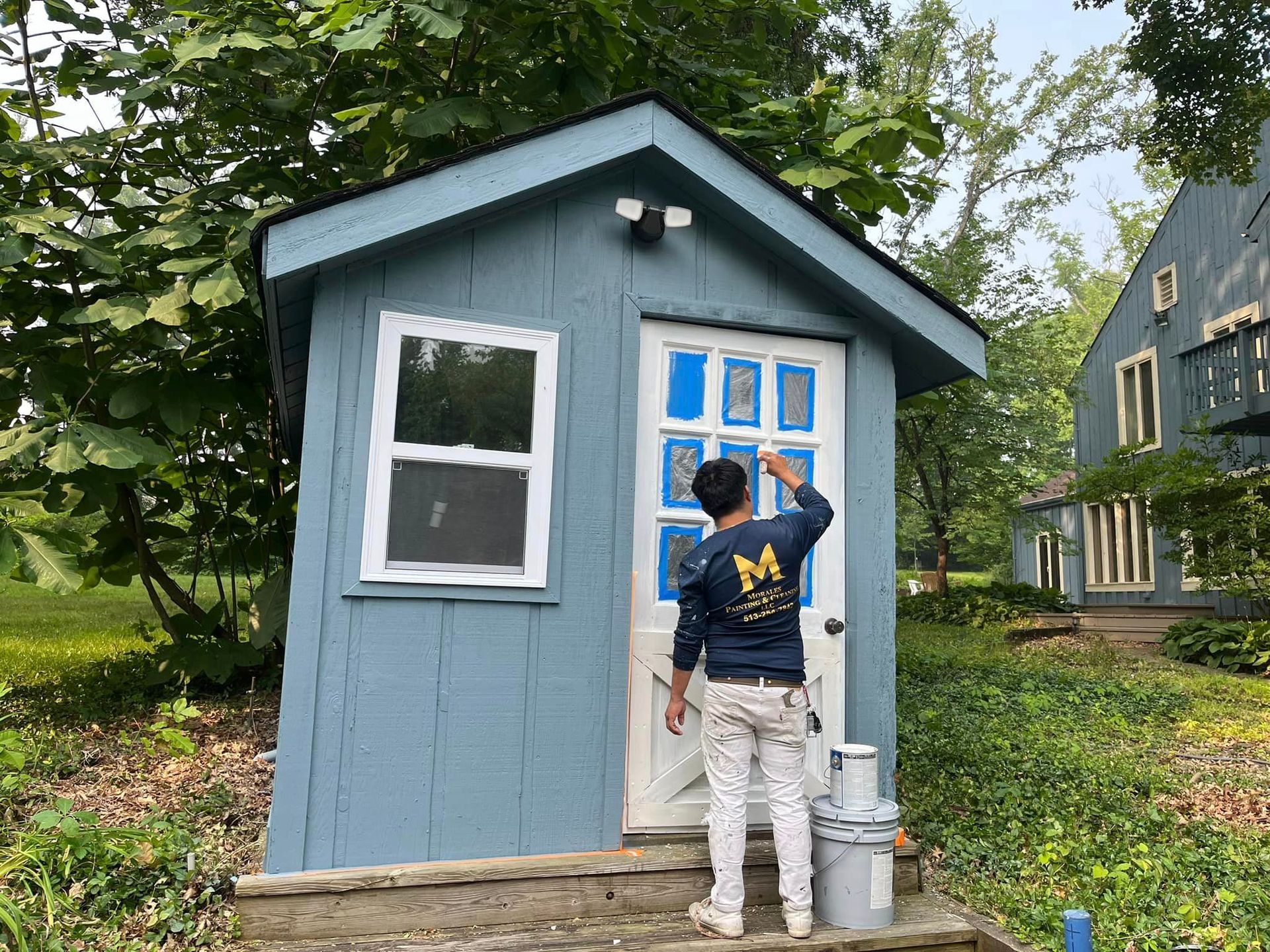 A man is painting the door of a blue shed.