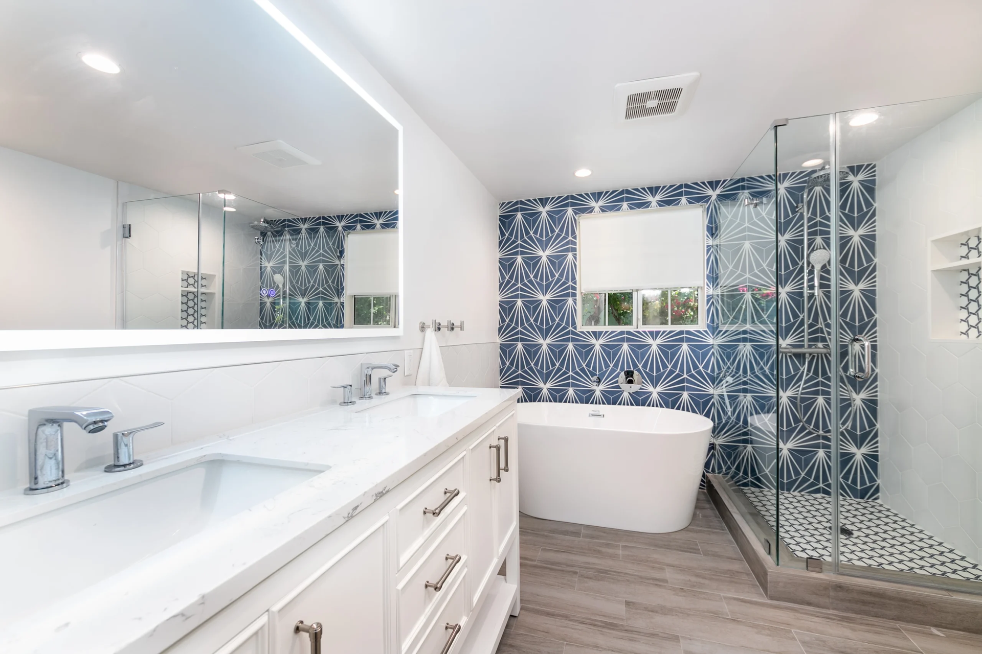 Modern bathroom with blue patterned accent wall, white vanity, freestanding tub, and glass shower.