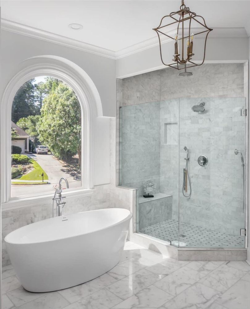 Elegant white bathroom with a soaking tub and walk-in shower. Large arched window and decorative light fixture.
