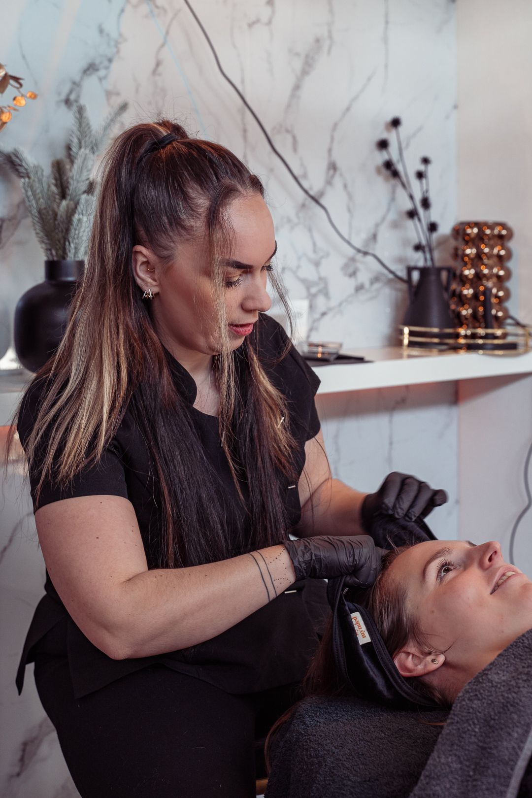 Een schoonheidsspecialist is bezig met het haar van een klant in een salon, met een marmeren muur en planken op de achtergrond.