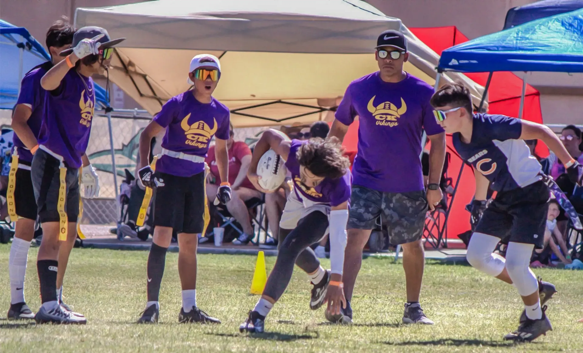 Youth sports players in purple jerseys crouch at a field start line under a canopy, ready to race