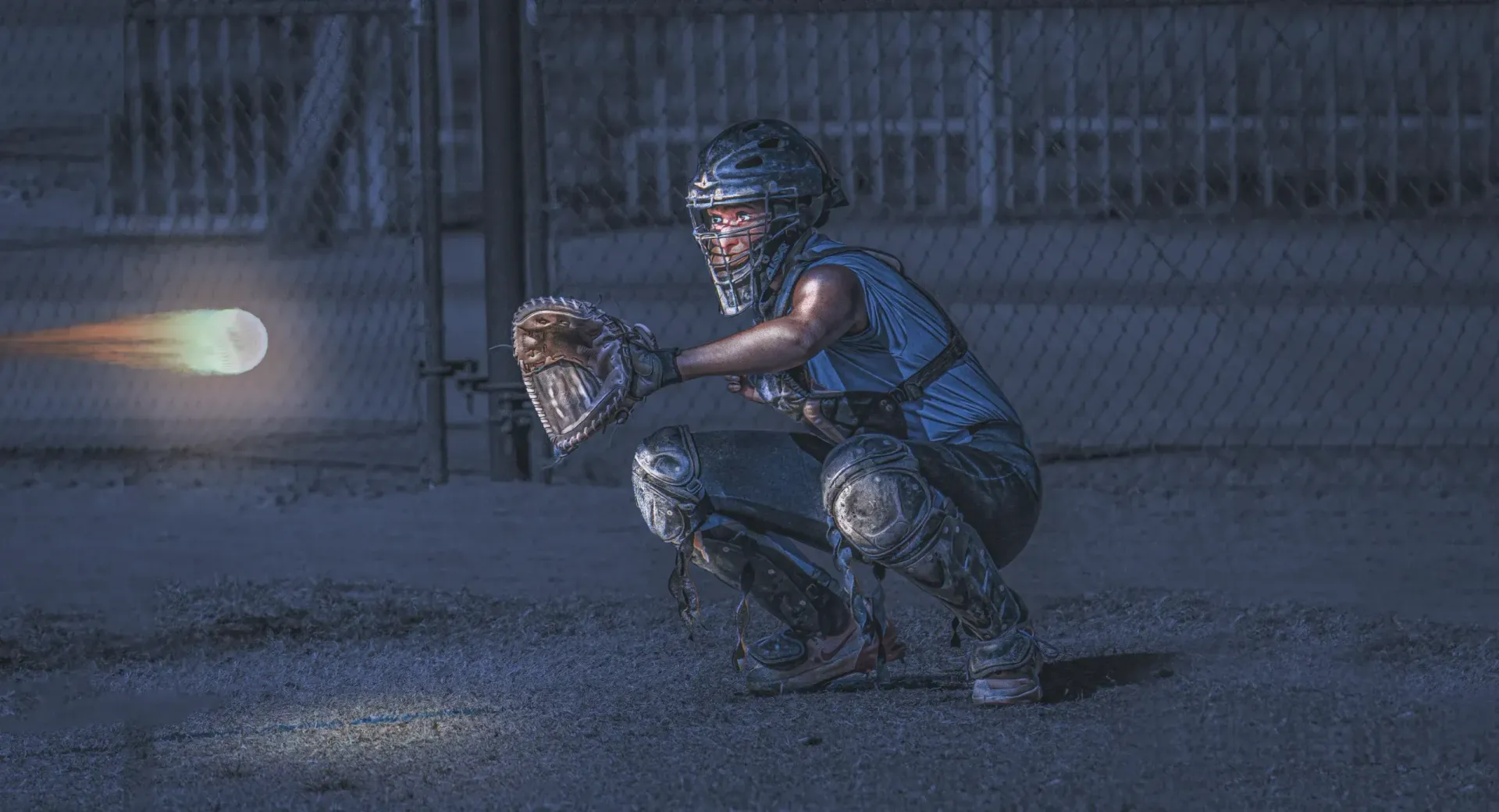 Baseball catcher crouching behind home plate as a pitch flies in at night