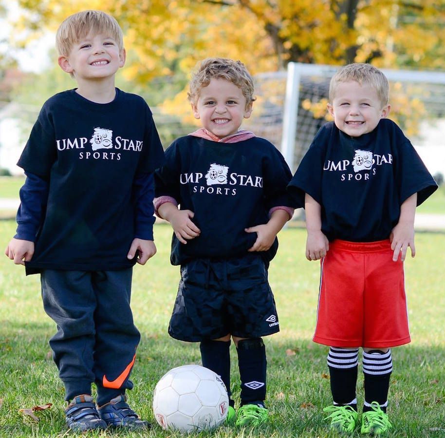 Three young boys wearing ump star sports shirts stand next to a soccer ball