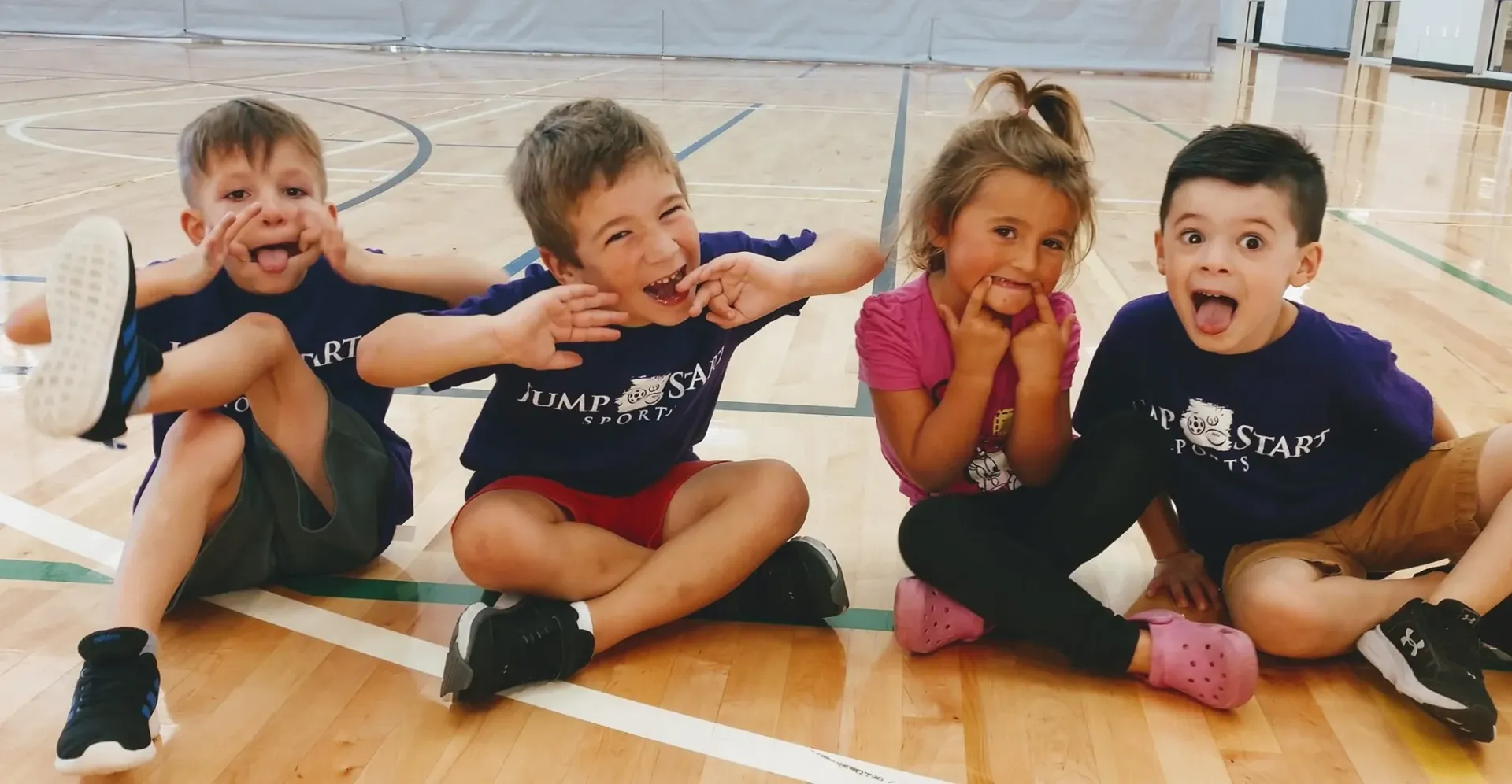 Four children are sitting on the floor in a gym making funny faces.