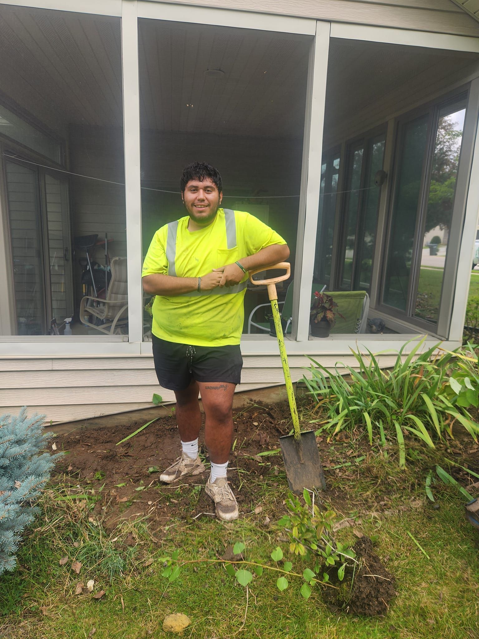 A man in a yellow shirt is standing in front of a house holding a shovel.