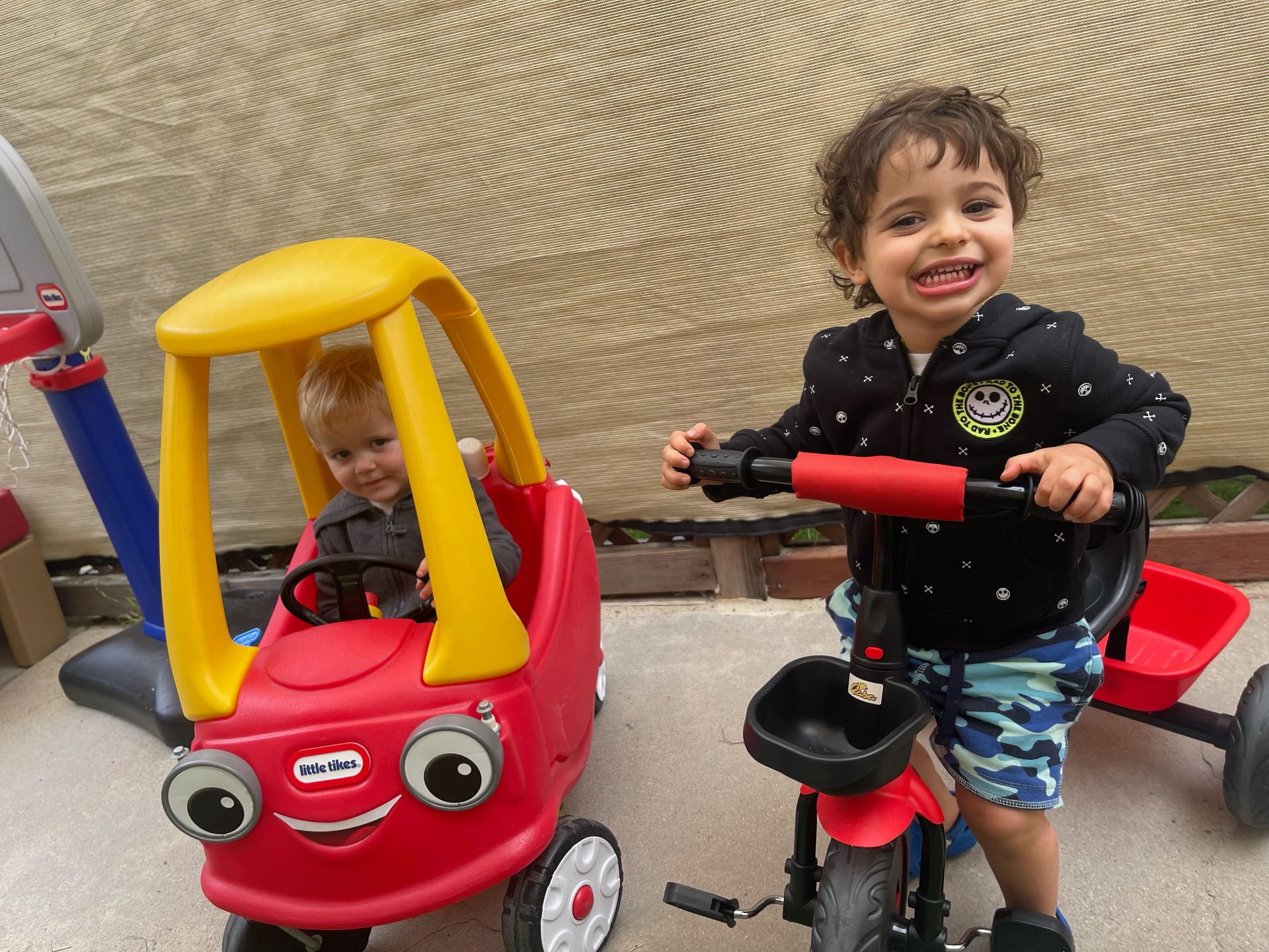 Two children smile near a red toy car and tricycle. One sits in the car, the other rides the trike, both outside.