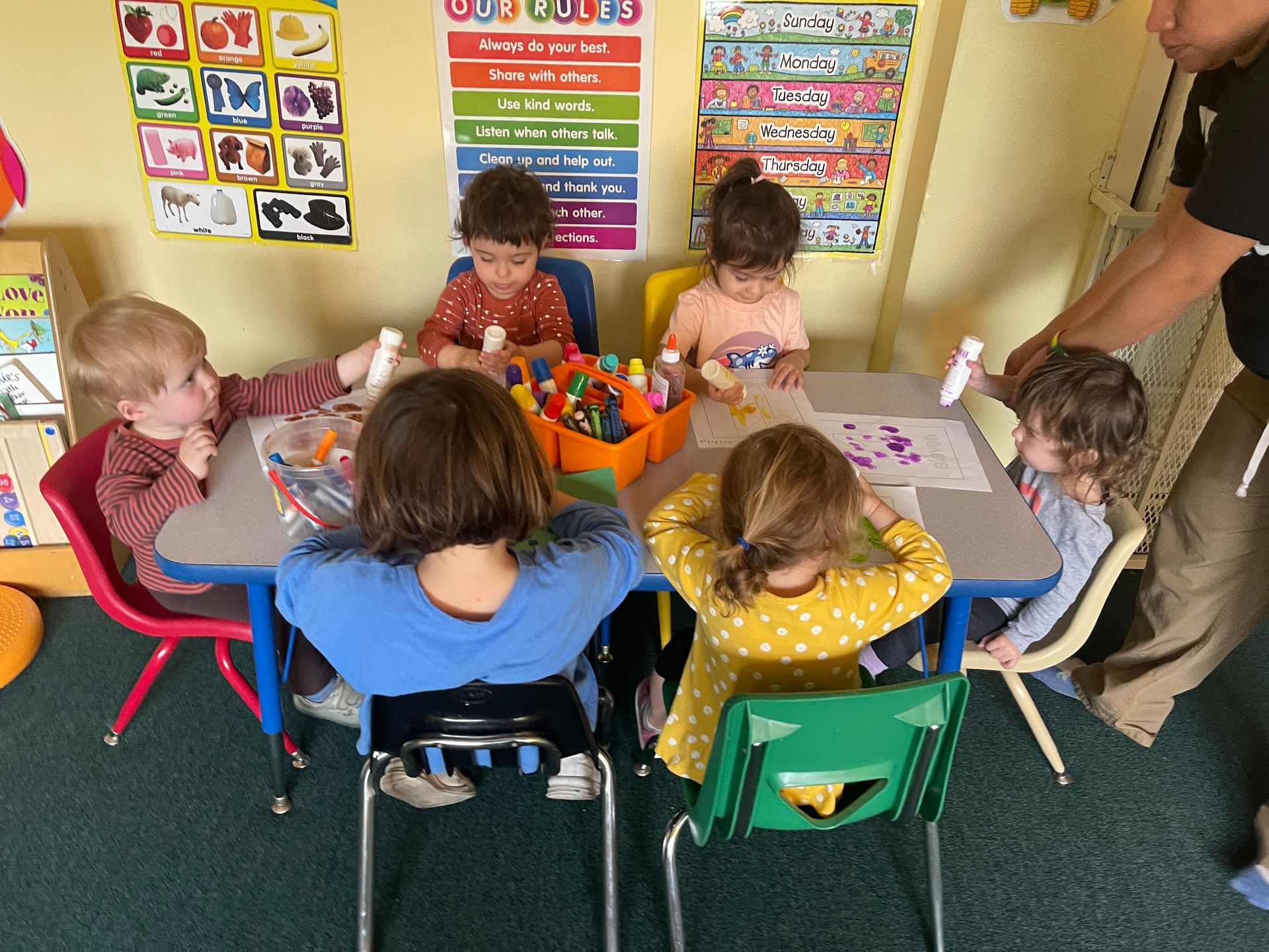 Children sitting at a table in a classroom, crafting. An adult is assisting.