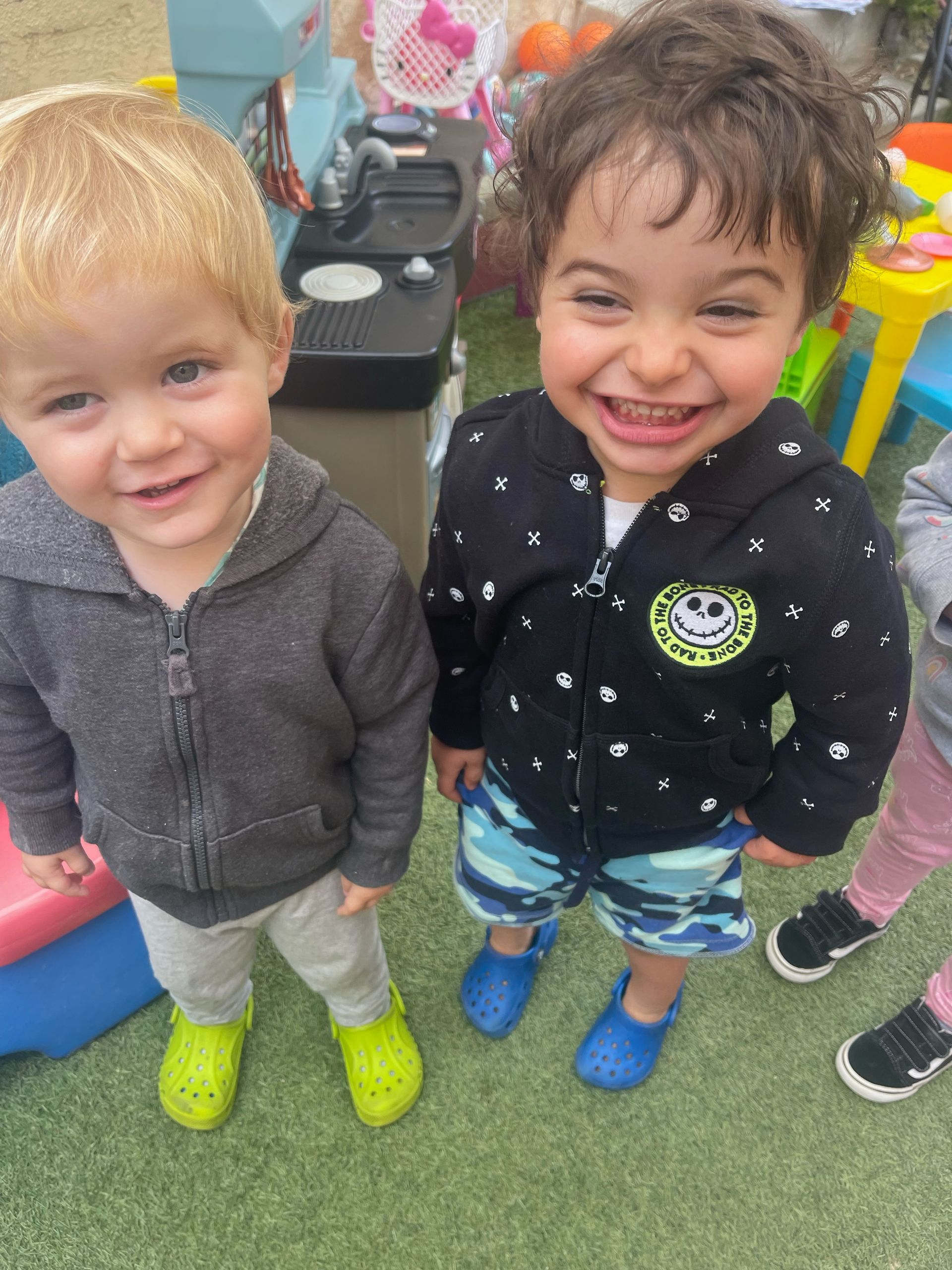 Two toddlers smiling, one in gray hoodie, green shoes, the other in black jacket, blue shorts, blue shoes. Outdoors.