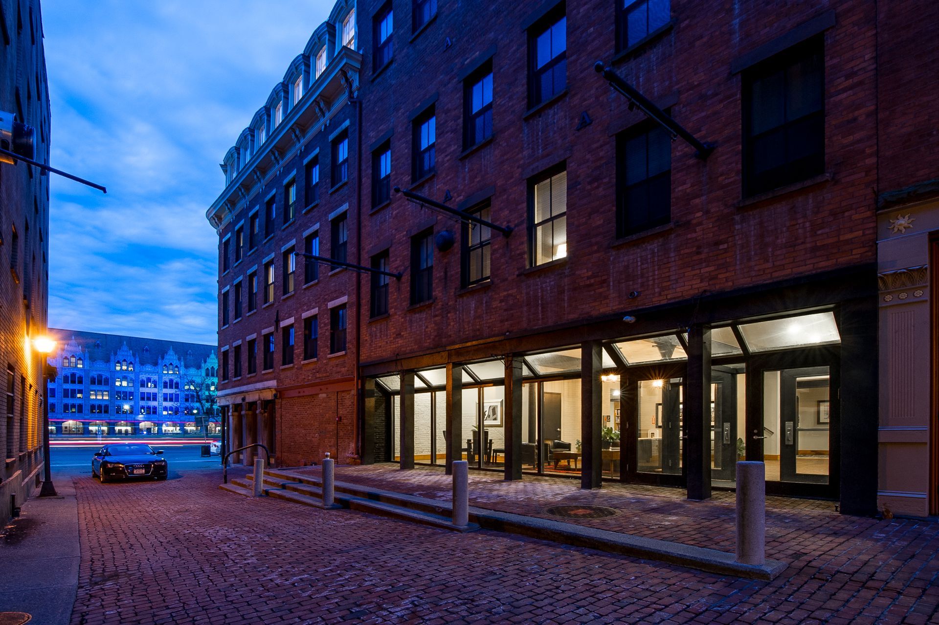 A brick building with a lot of windows is next to a cobblestone street.