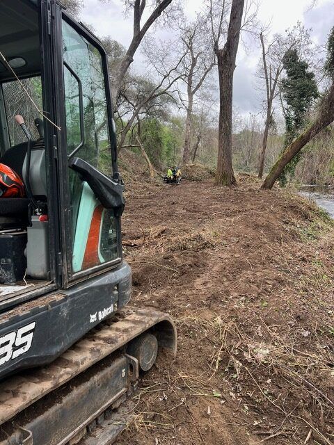 A small excavator is sitting on top of a dirt hill next to a river.