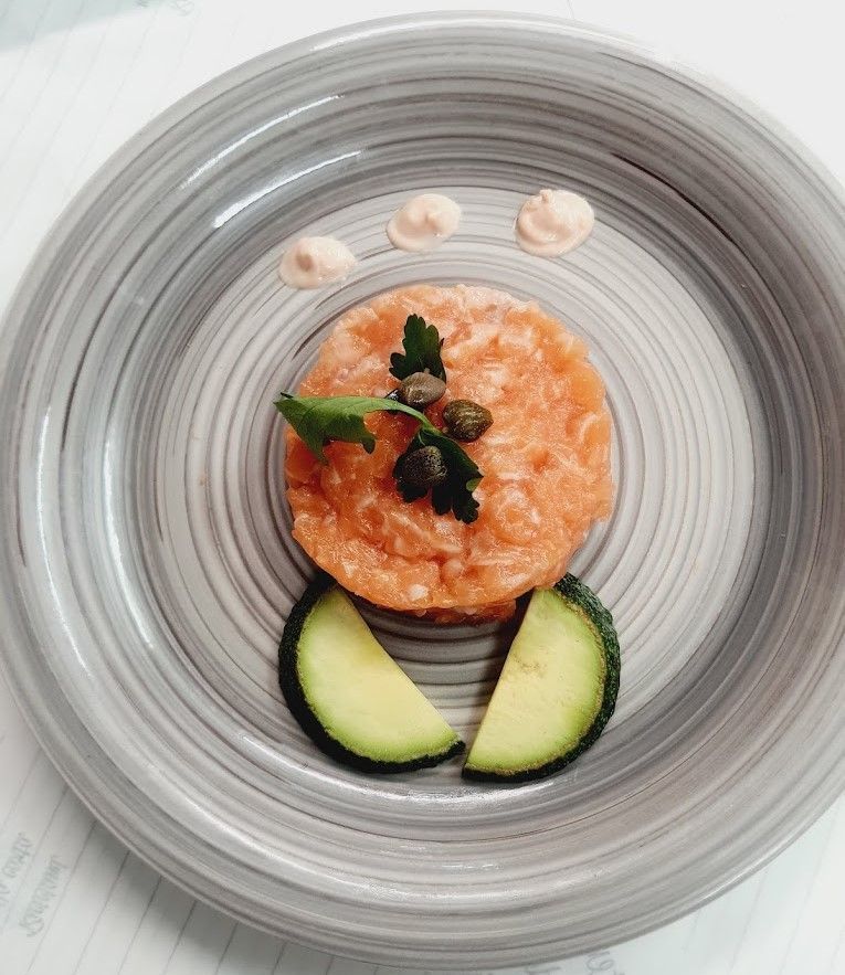 Tartare di salmone con capperi, prezzemolo, salsa e fette di zucchine su un piatto grigio.