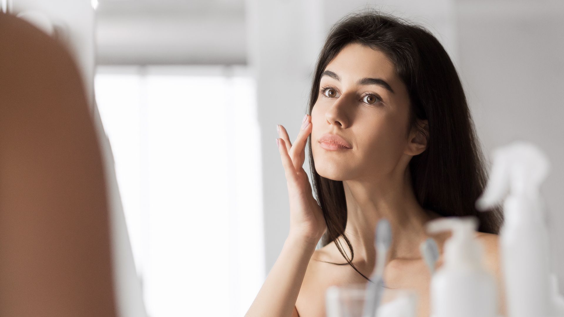 A person looking into a mirror while gently touching their cheek, standing in a bright, modern bathroom.