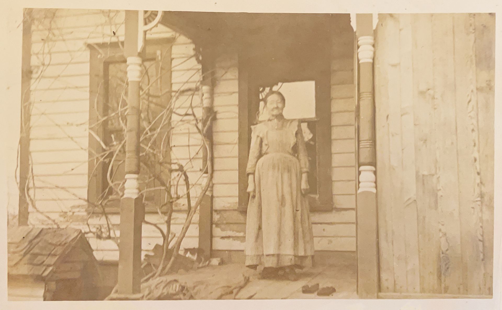 A woman in a long dress is standing on the porch of a house.