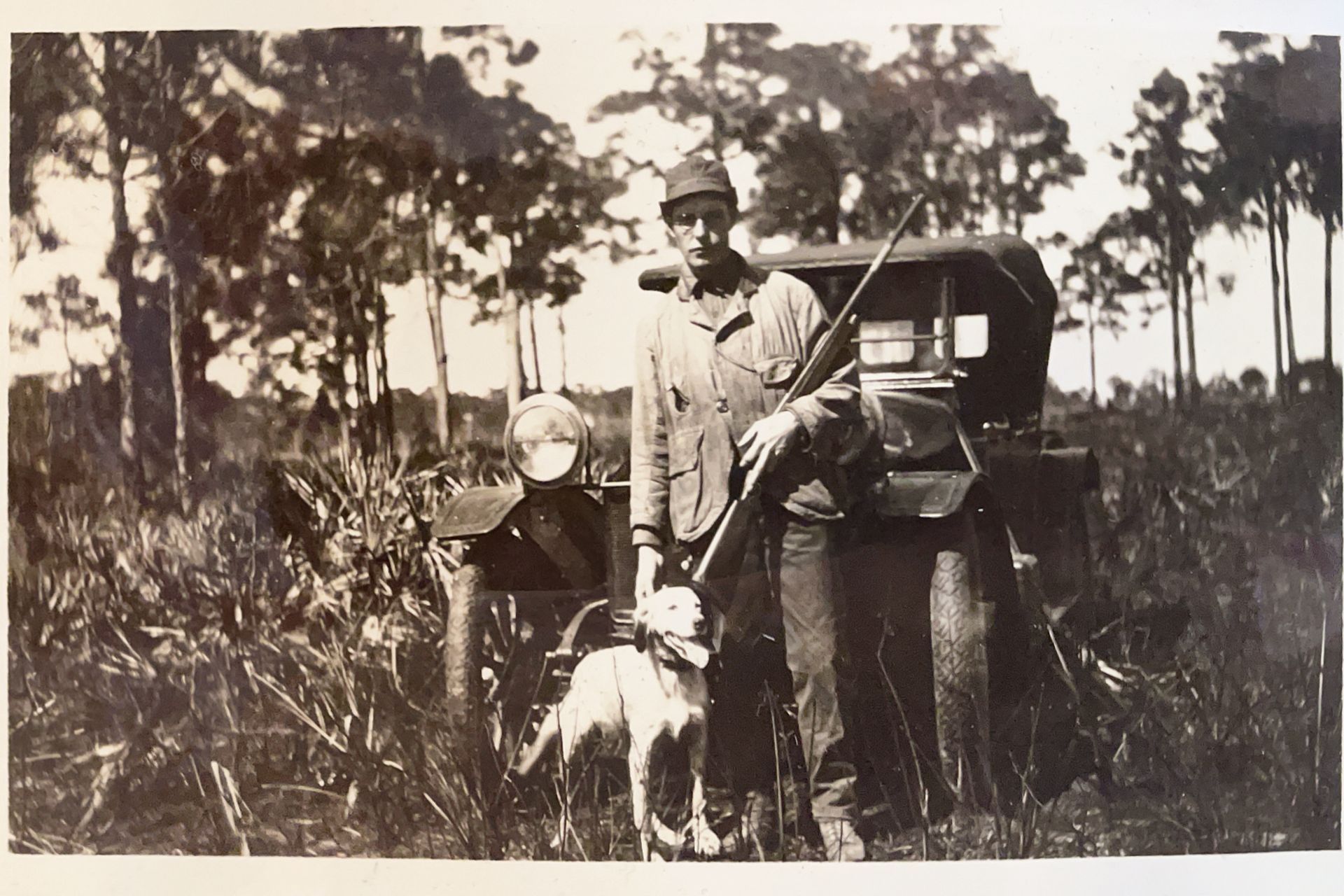 historical picture of man holding rifle, standing with a dog, in front of a motor vehicle