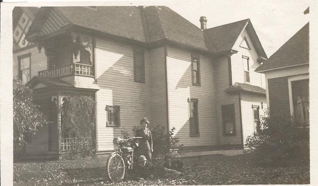 A black and white photo of a man sitting on a motorcycle in front of a house