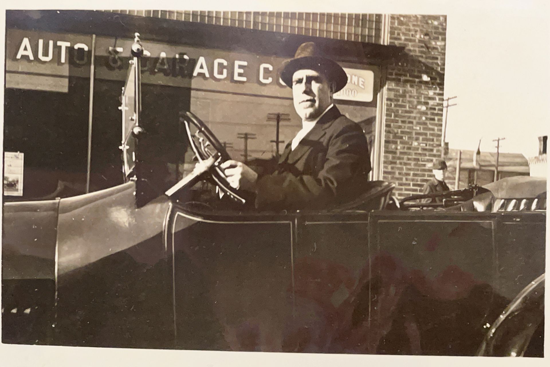 A black and white photo of a man in a car in front of an auto garage