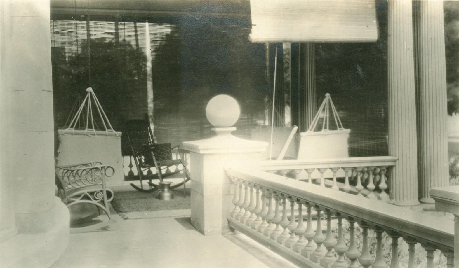 A black and white photo of a porch with a railing and chairs.