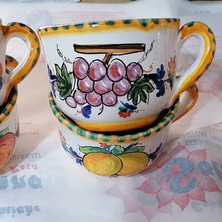 Due tazze di ceramica colorate, una sopra l'altra, con motivi di uva e frutta, finiture gialle e manici.