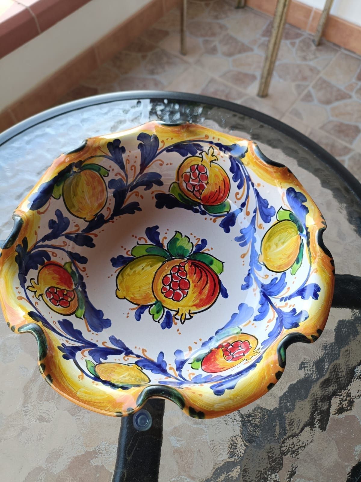 Ciotola in ceramica con motivi di frutta gialla, blu e arancione su un tavolo di vetro.