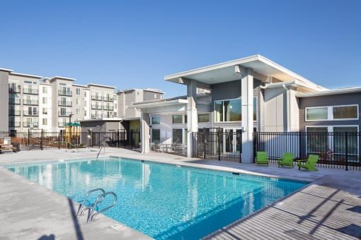 Swimming pool in front of an apartment complex. Gray and white buildings, green chairs, sunny day.