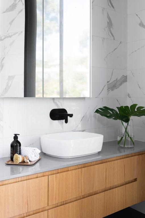 Modern bathroom with white vessel sink, black faucet, wooden cabinet, marble tile, and a vase with a monstera leaf.