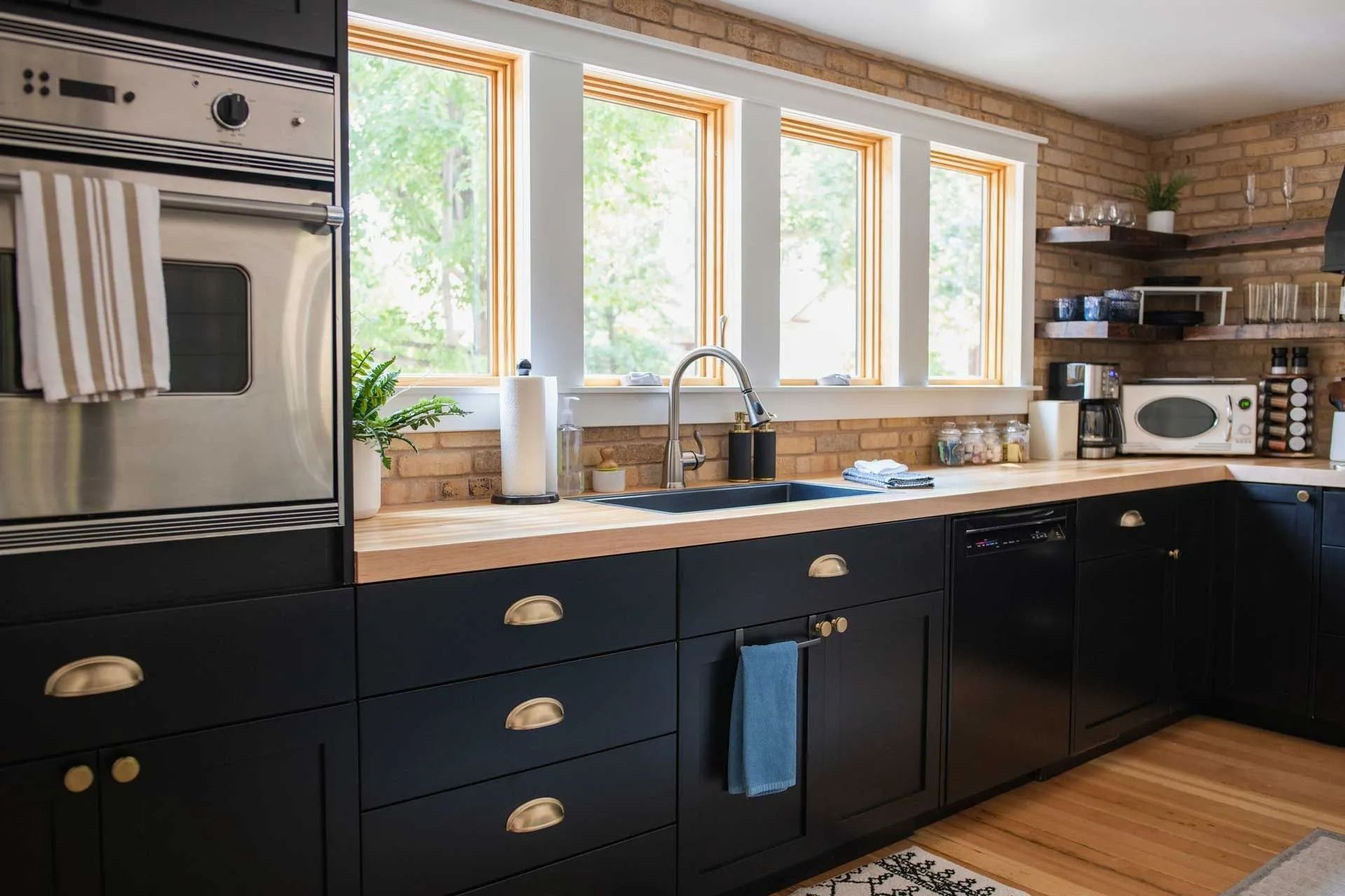 Black kitchen cabinets with brass hardware, wooden countertop, and sink under three windows. Black kitchen cabinets with brass hardware, wooden countertop, and sink under three windows.