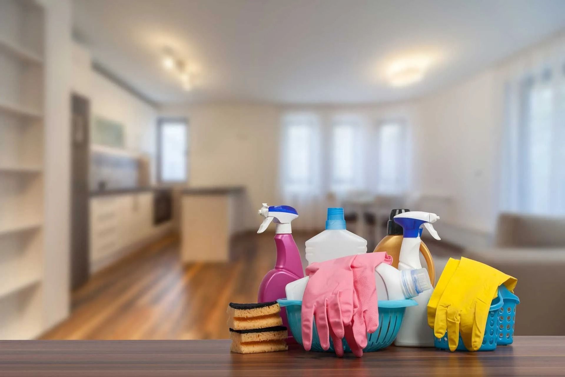Cleaning supplies in front of a blurred living room: spray bottles, gloves, sponges, and a bucket.