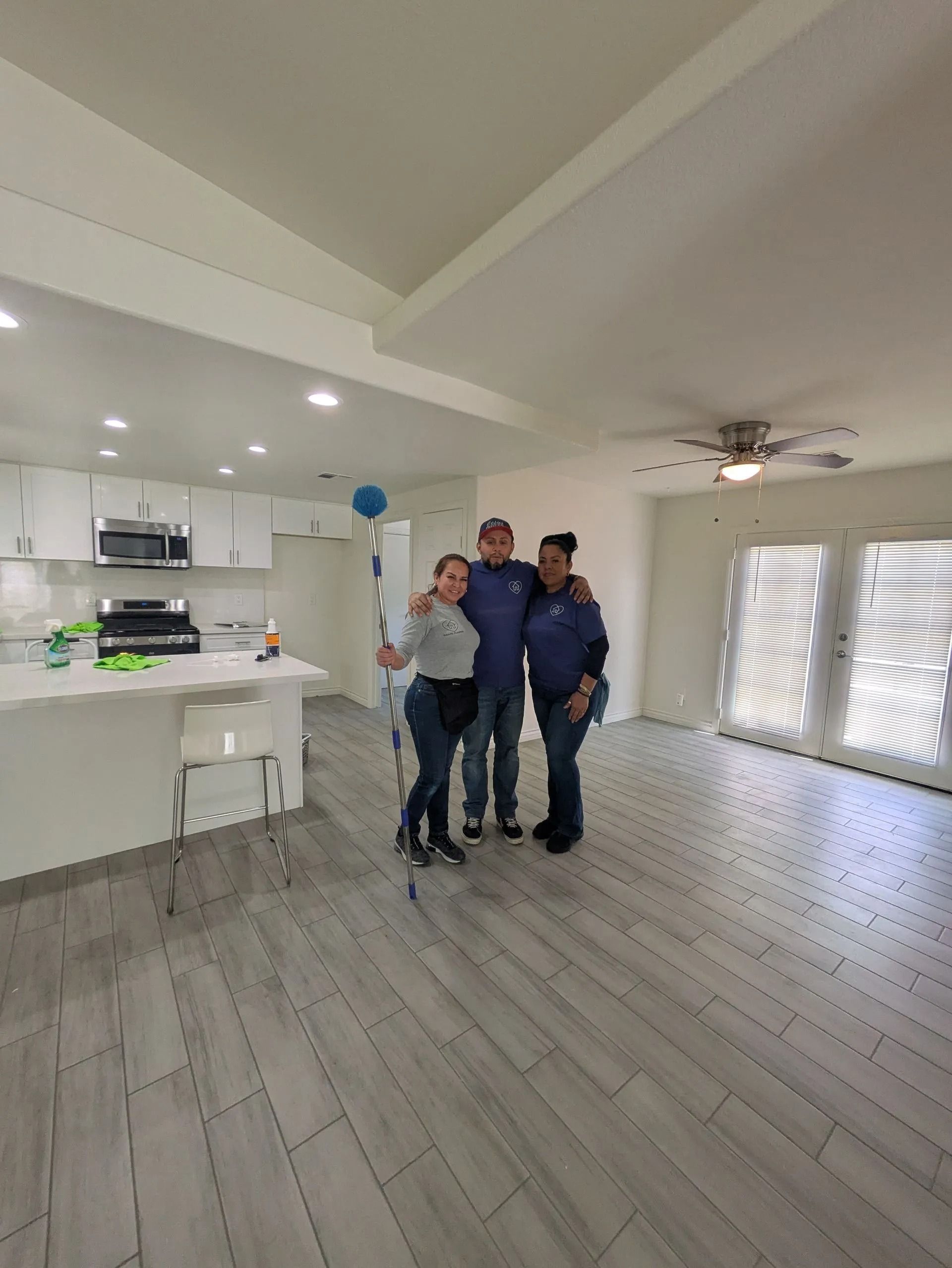 Three people pose in a bright, newly renovated room. White cabinets, gray floors, and French doors are visible. Three people pose in a bright, newly renovated room. White cabinets, gray floors, and French doors are visible.
