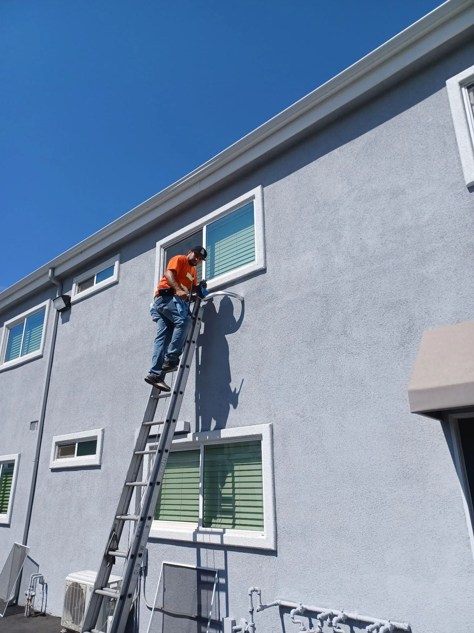 Person on ladder, working on a window of a light gray building against a blue sky.