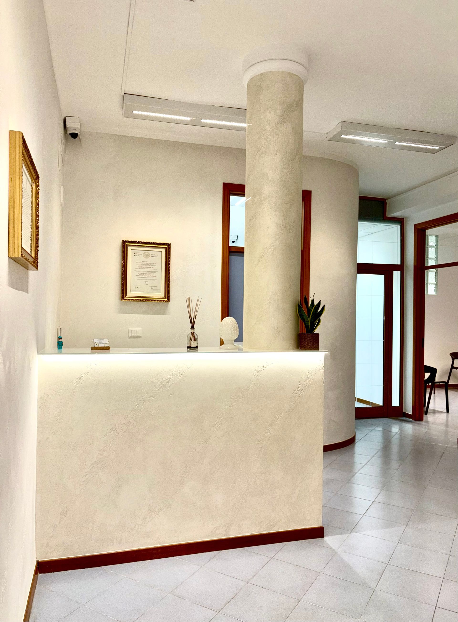 Un bancone reception color crema si trova nella hall di un ufficio luminoso e moderno, caratterizzato da una colonna centrale e da opere d'arte incorniciate alle pareti.