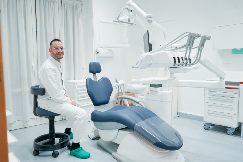 Un professionista è seduto su uno sgabello con le rotelle accanto a una poltrona odontoiatrica blu in una clinica luminosa e moderna.