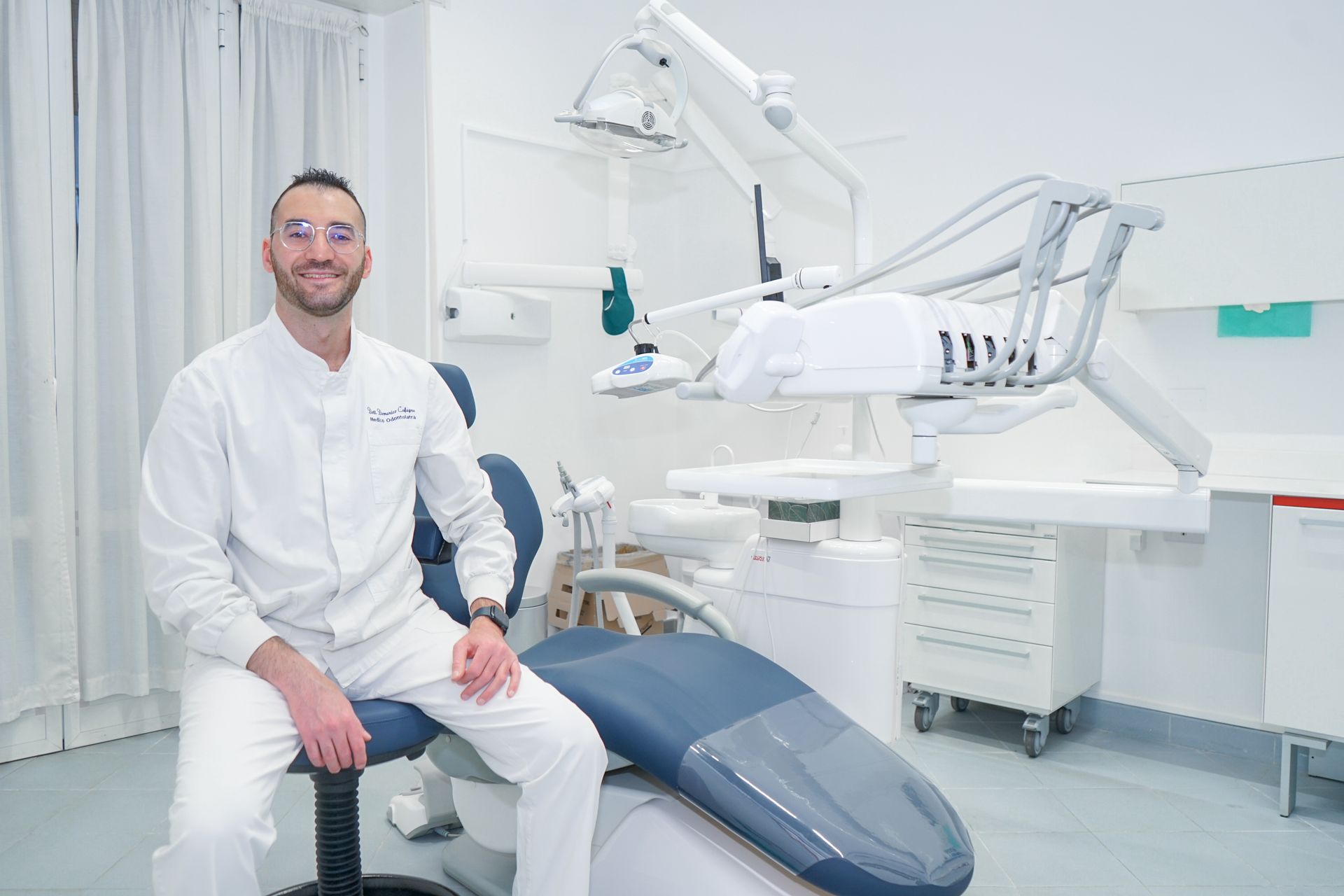 In una clinica luminosa e moderna, un dentista in camice bianco e occhiali protettivi siede accanto a una poltrona odontoiatrica.