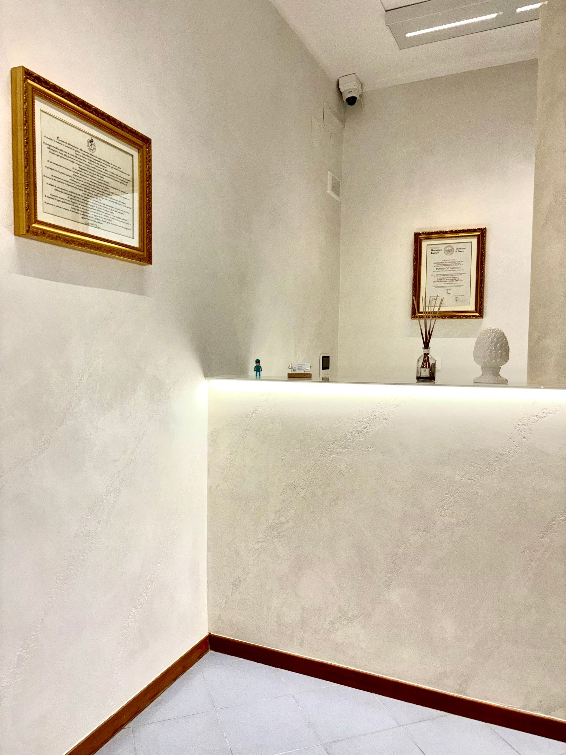 Una reception minimalista con pareti color avorio materico, un bancone illuminato, due certificati incorniciati e una piccola scultura.