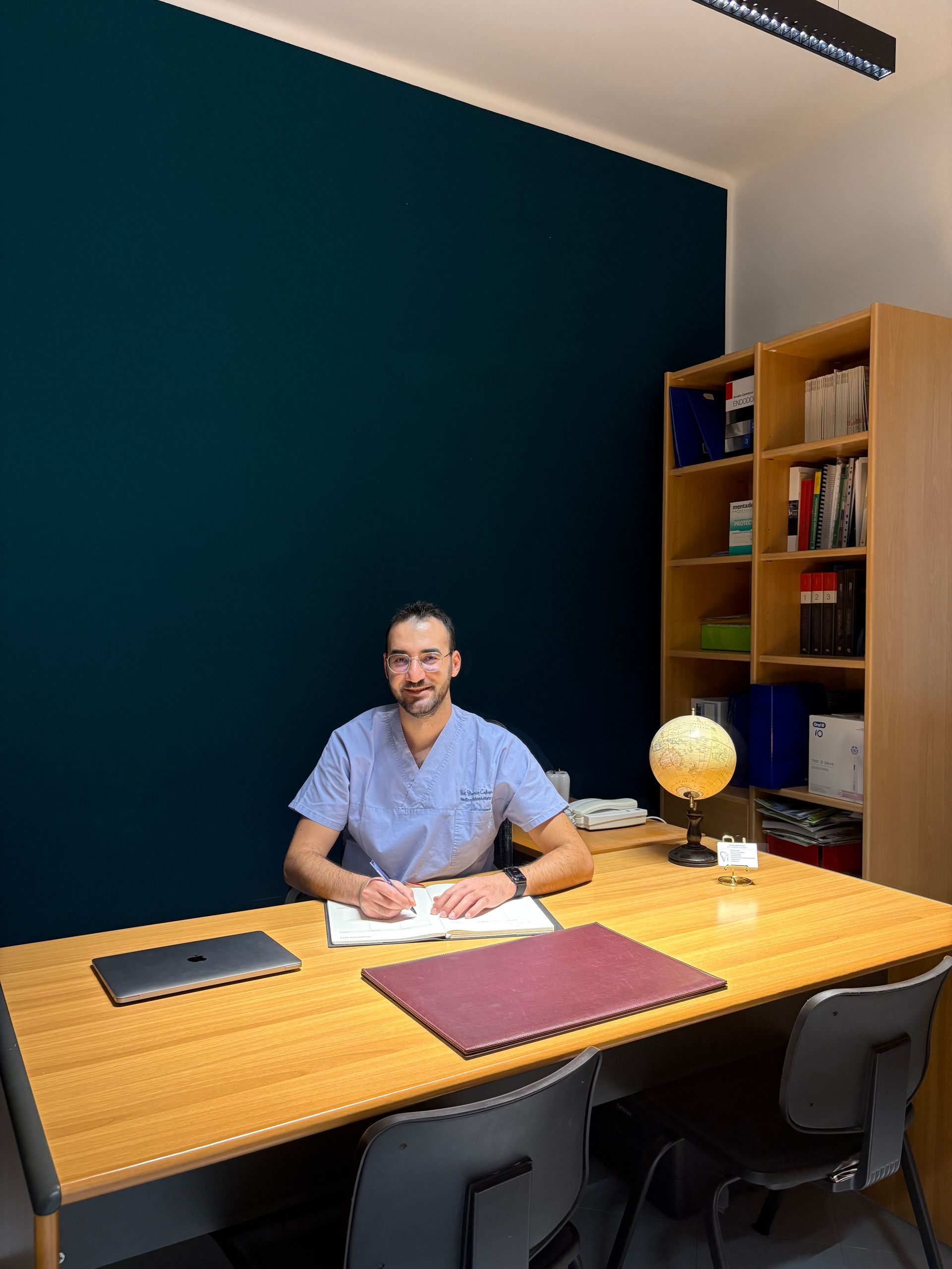 Una persona in camice blu è seduta a una scrivania in ufficio, intenta a scrivere su un quaderno accanto a un computer portatile, un mappamondo e una libreria di legno.