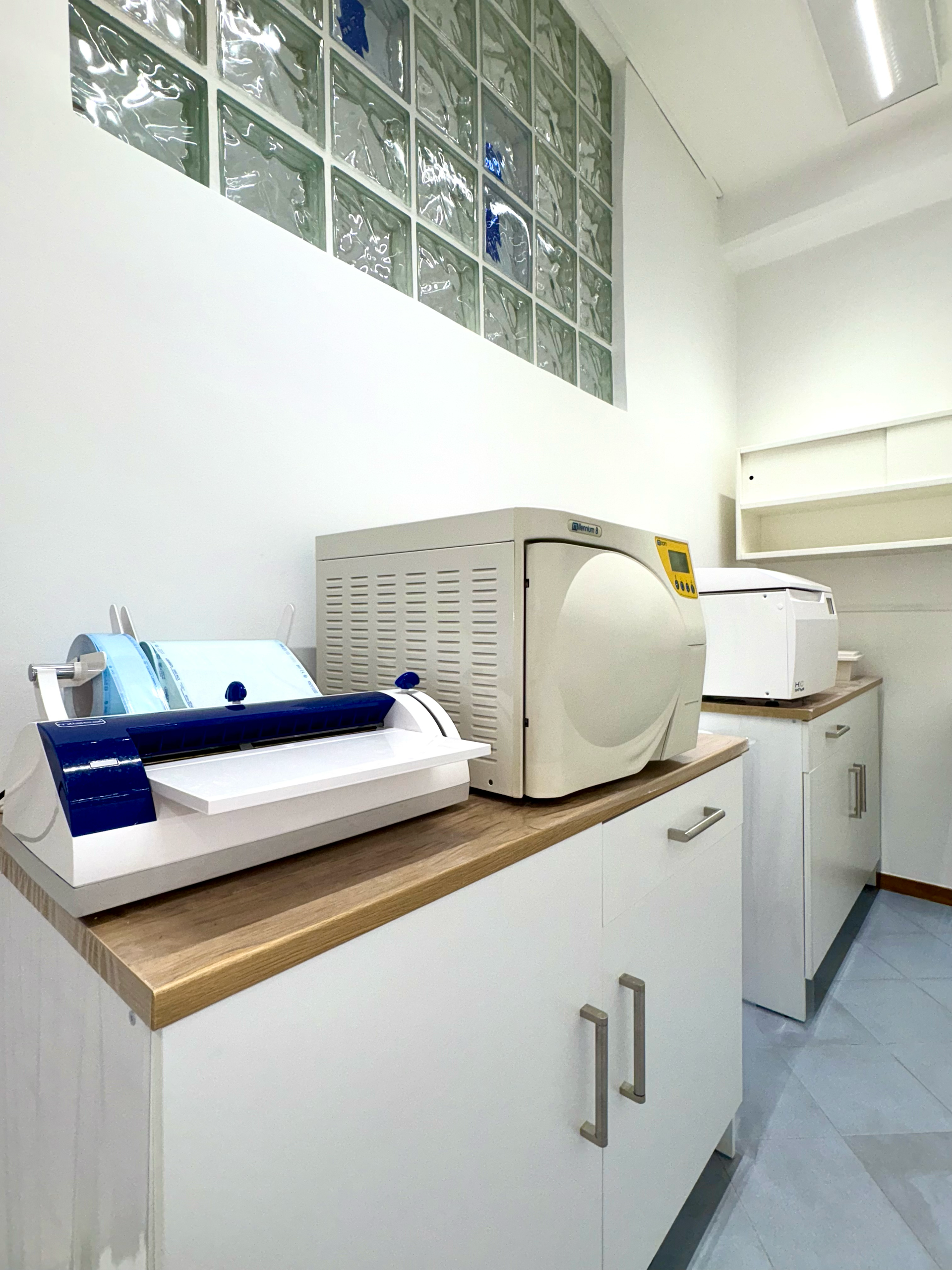 Una sala di sterilizzazione medicale dotata di una sigillatrice per buste e di un'autoclave da banco, entrambe collocate su armadietti bianchi sotto blocchi di vetro.