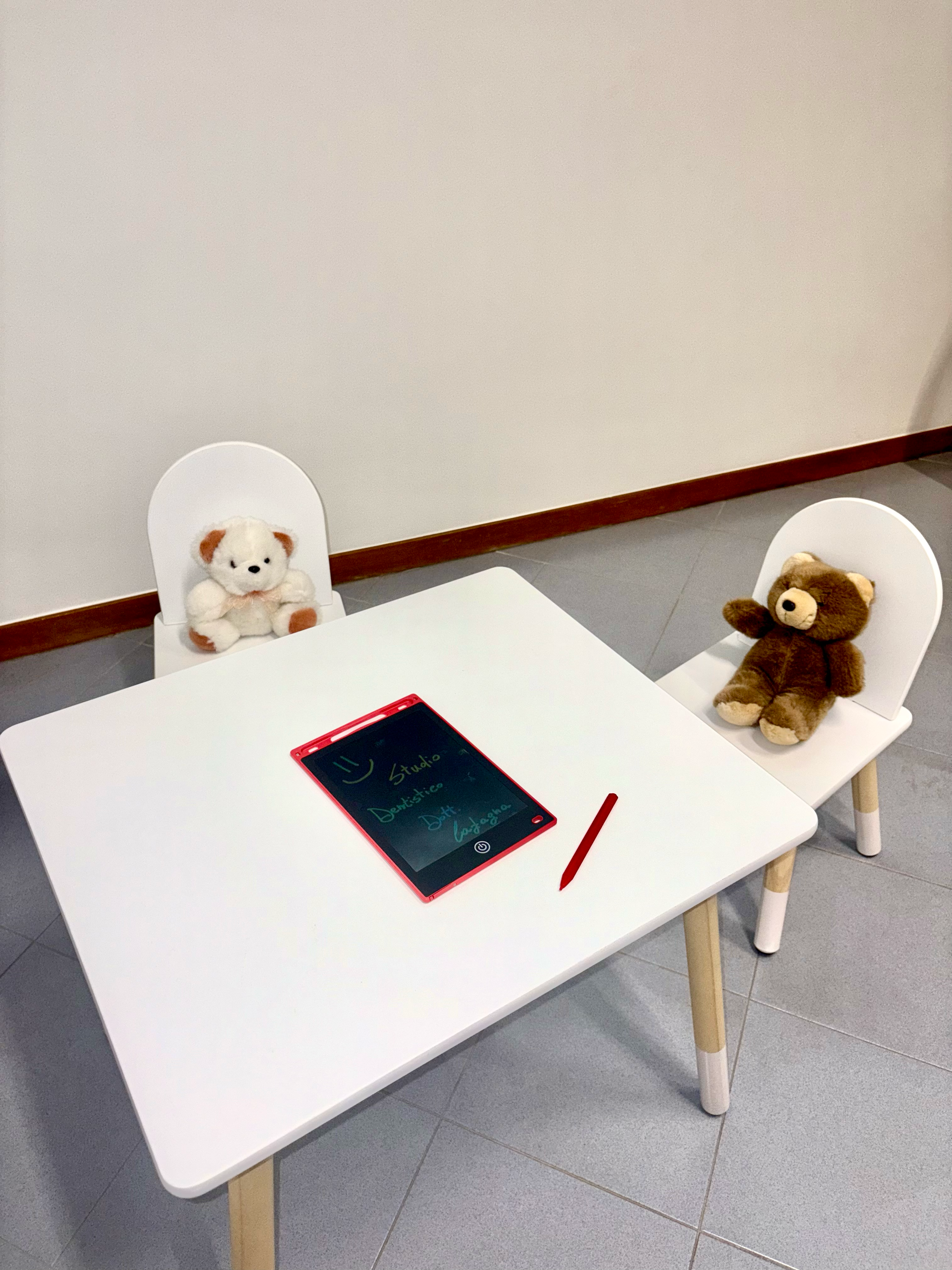 Un tavolino bianco per bambini con due piccole sedie, ognuna con un orsacchiotto di peluche, e al centro un tablet per scrivere con stilo.