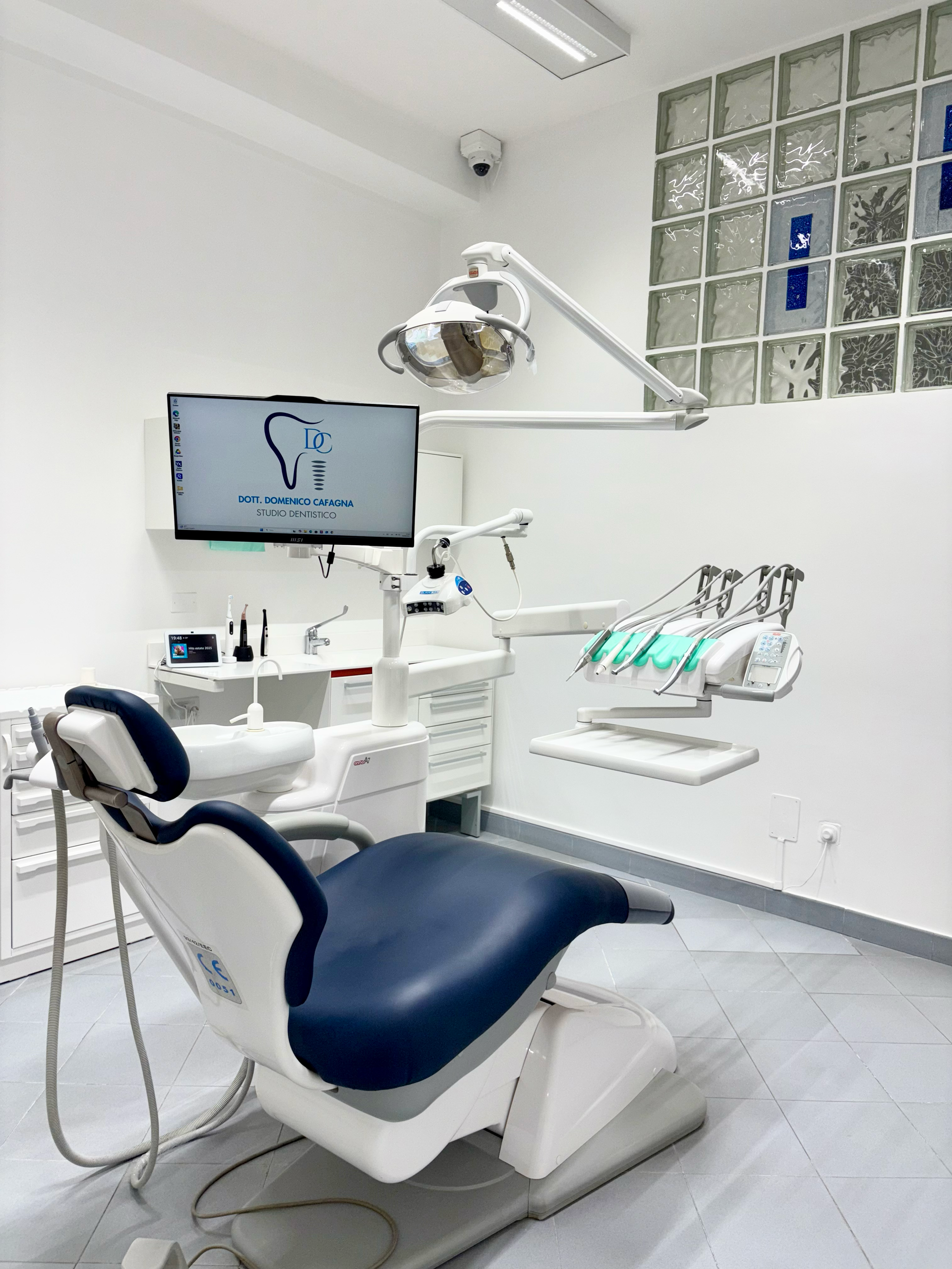 Una sala per visite odontoiatriche con una poltrona bianca, cuscini blu, attrezzature mediche specializzate e un monitor su un supporto.