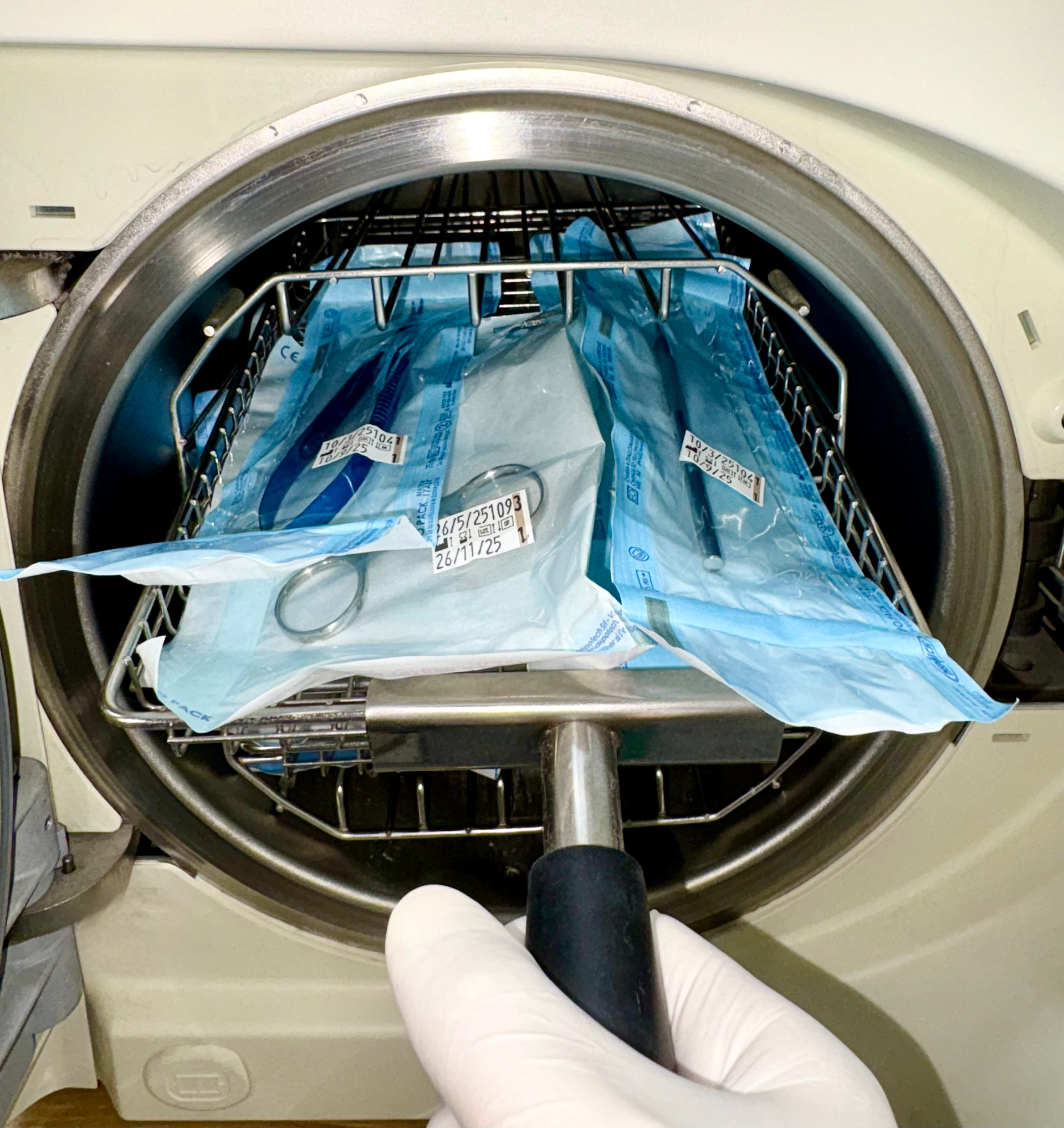 Una mano guantata fa scivolare un vassoio metallico contenente strumenti odontoiatrici avvolti in pellicola sterile all'interno di un'autoclave sterilizzatrice.
