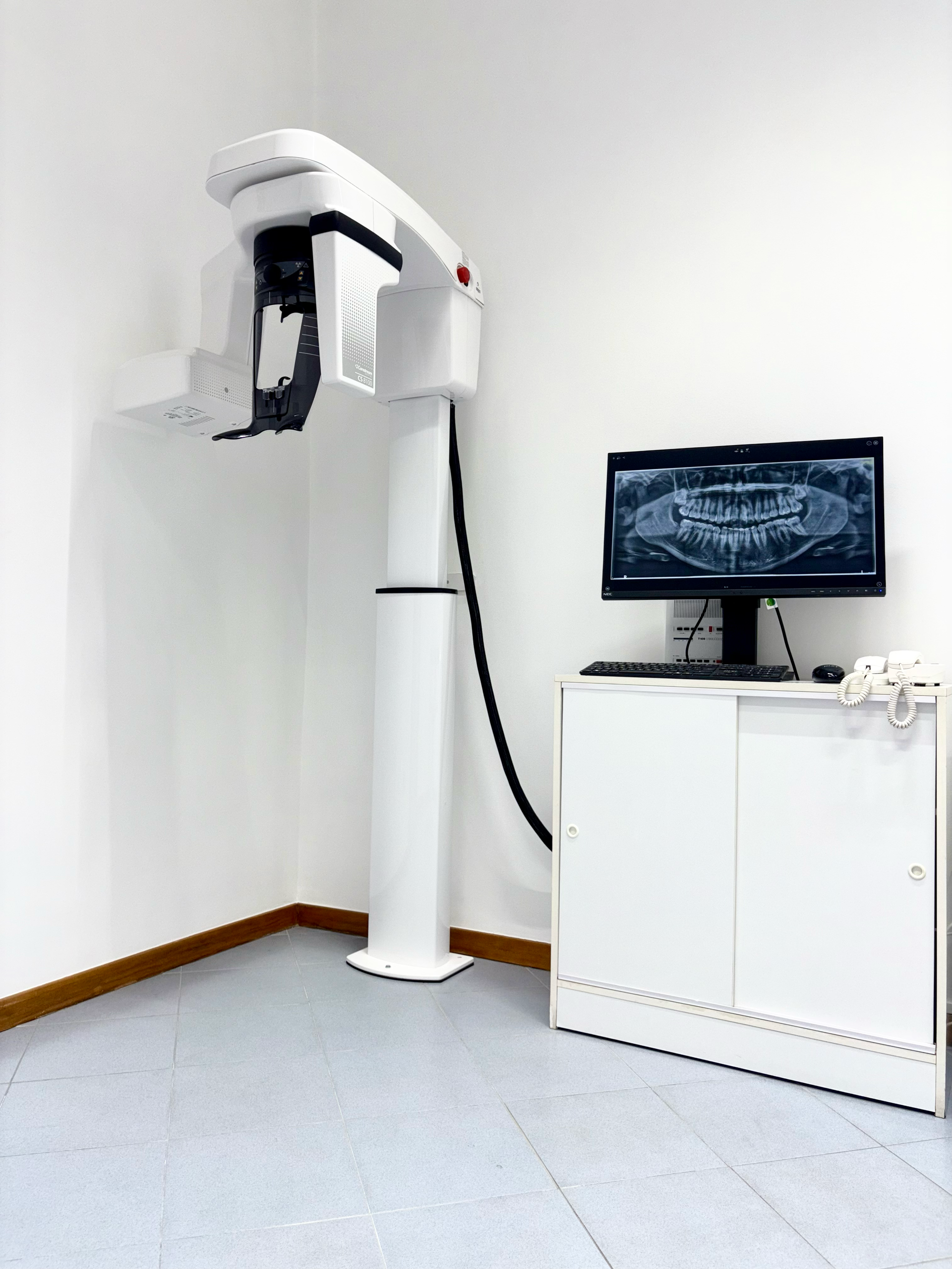 In una stanza bianca, un apparecchio radiografico dentale è posizionato accanto a un monitor che visualizza una scansione panoramica della colonna vertebrale.