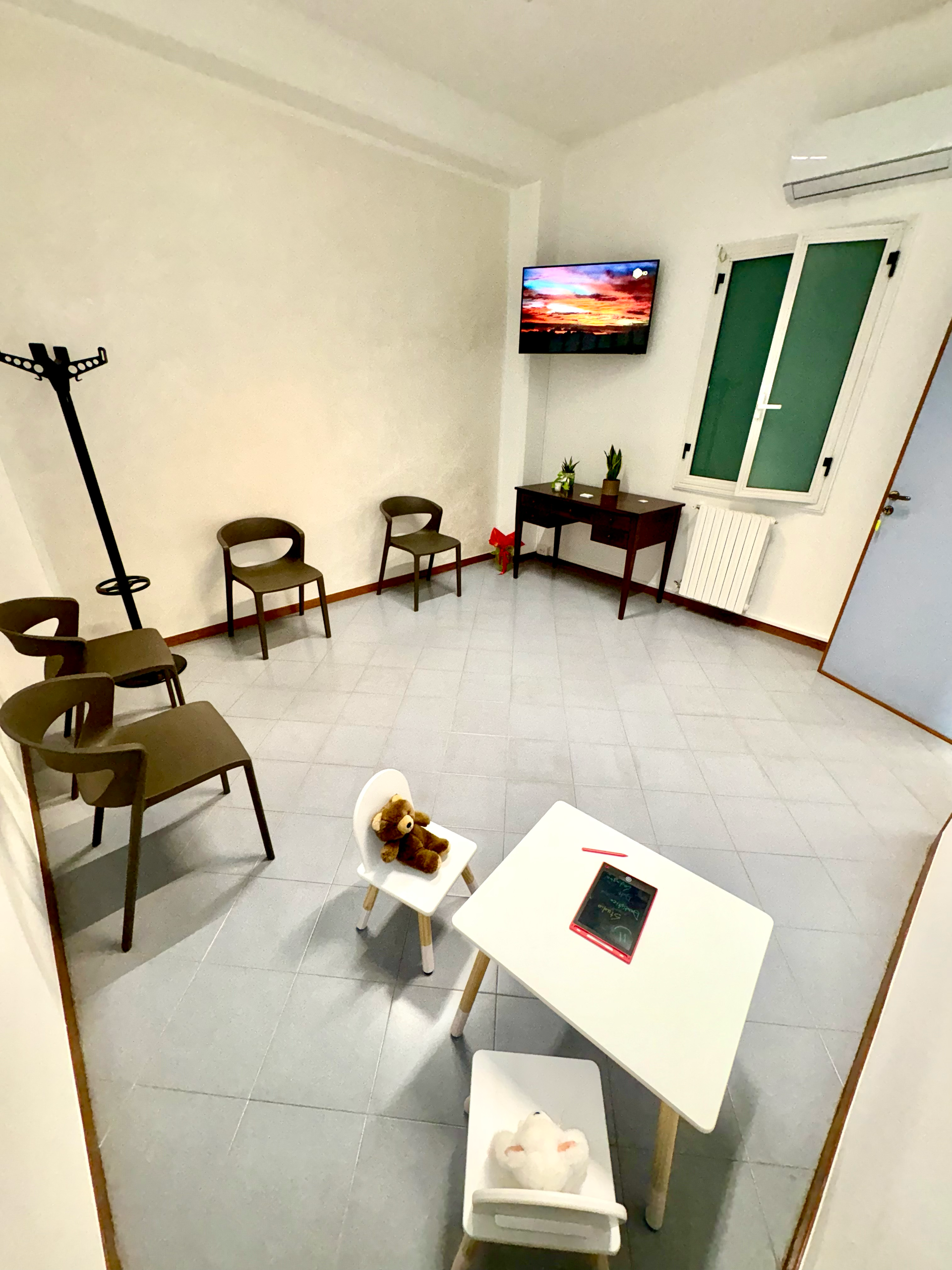 Una sala d'attesa con sedie disposte lungo la parete, un tavolino per bambini, due orsetti di peluche e un televisore montato a parete.