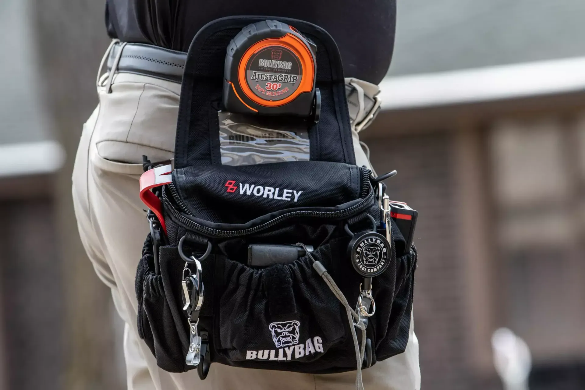 Person wearing a black tool belt, containing tape measure and other tools, attached to light-colored pants.