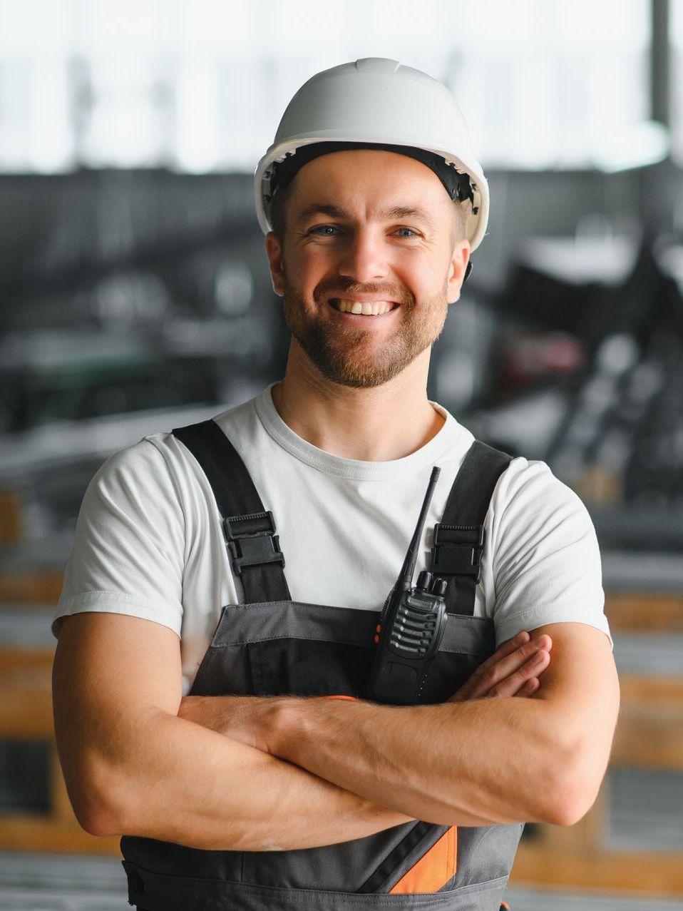 A man wearing a hard hat and overalls is smiling with his arms crossed.