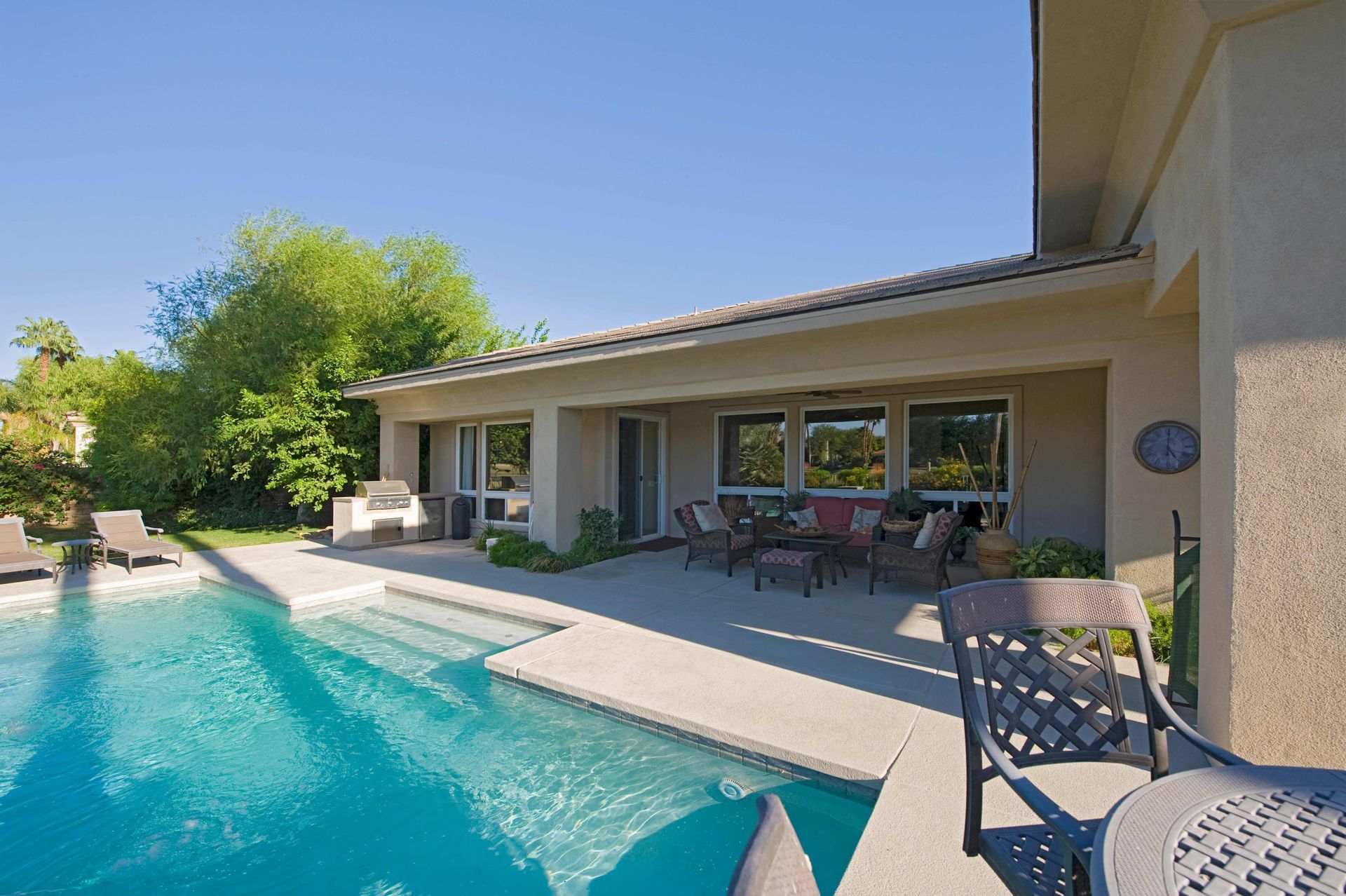 Backyard pool beside a covered patio with outdoor seating and a house, in bright sunlight