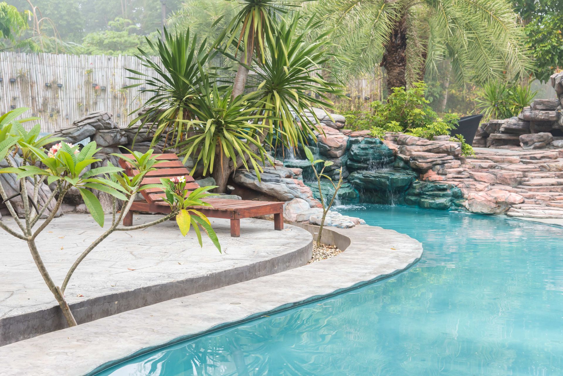 Curved turquoise pool with stone edging, tropical plants, and a small waterfall in a lush garden.