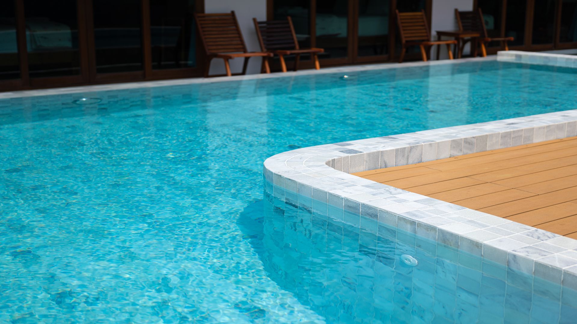 A clear blue swimming pool with a wooden deck area and lounge chairs in the background.