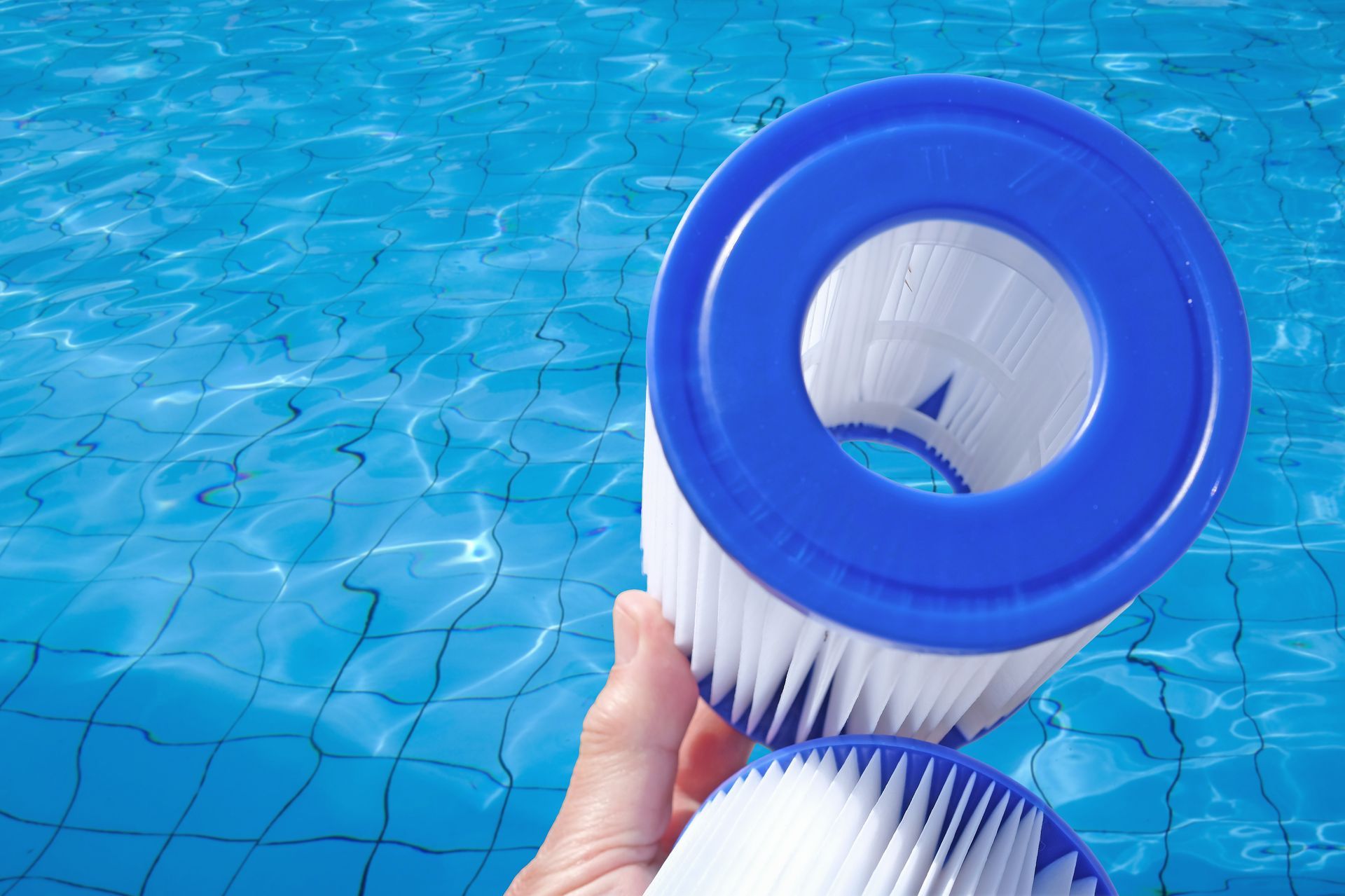 A hand holds a clean, pleated blue and white circular swimming pool filter over sparkling blue water.