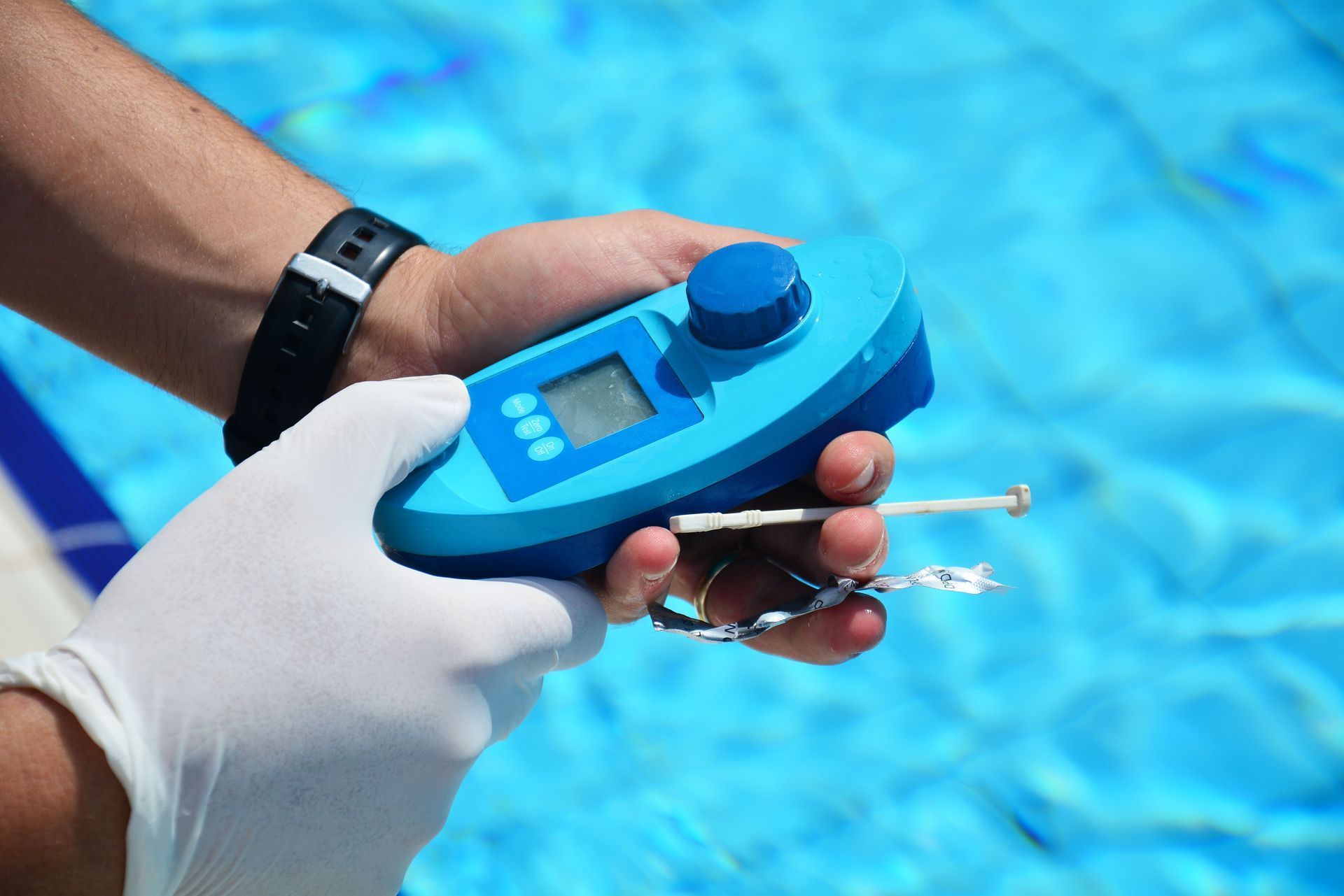 A gloved hand holds a blue electronic pool water tester device against a backdrop of rippling swimming pool water.